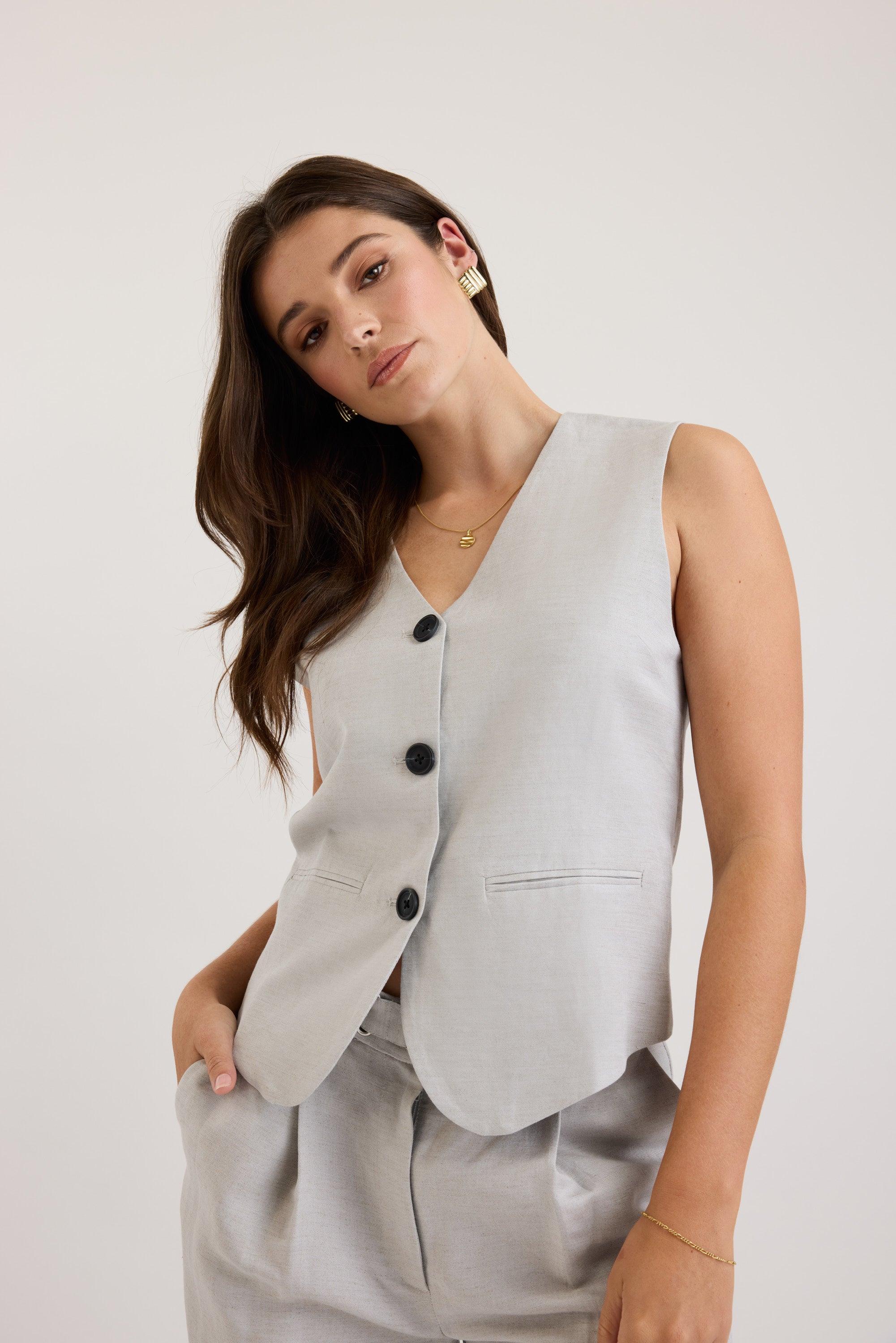 A woman with long brown hair models The Tailored Vest and matching pants by Max, accessorized with gold jewelry, standing against a plain light backdrop with a relaxed expression.