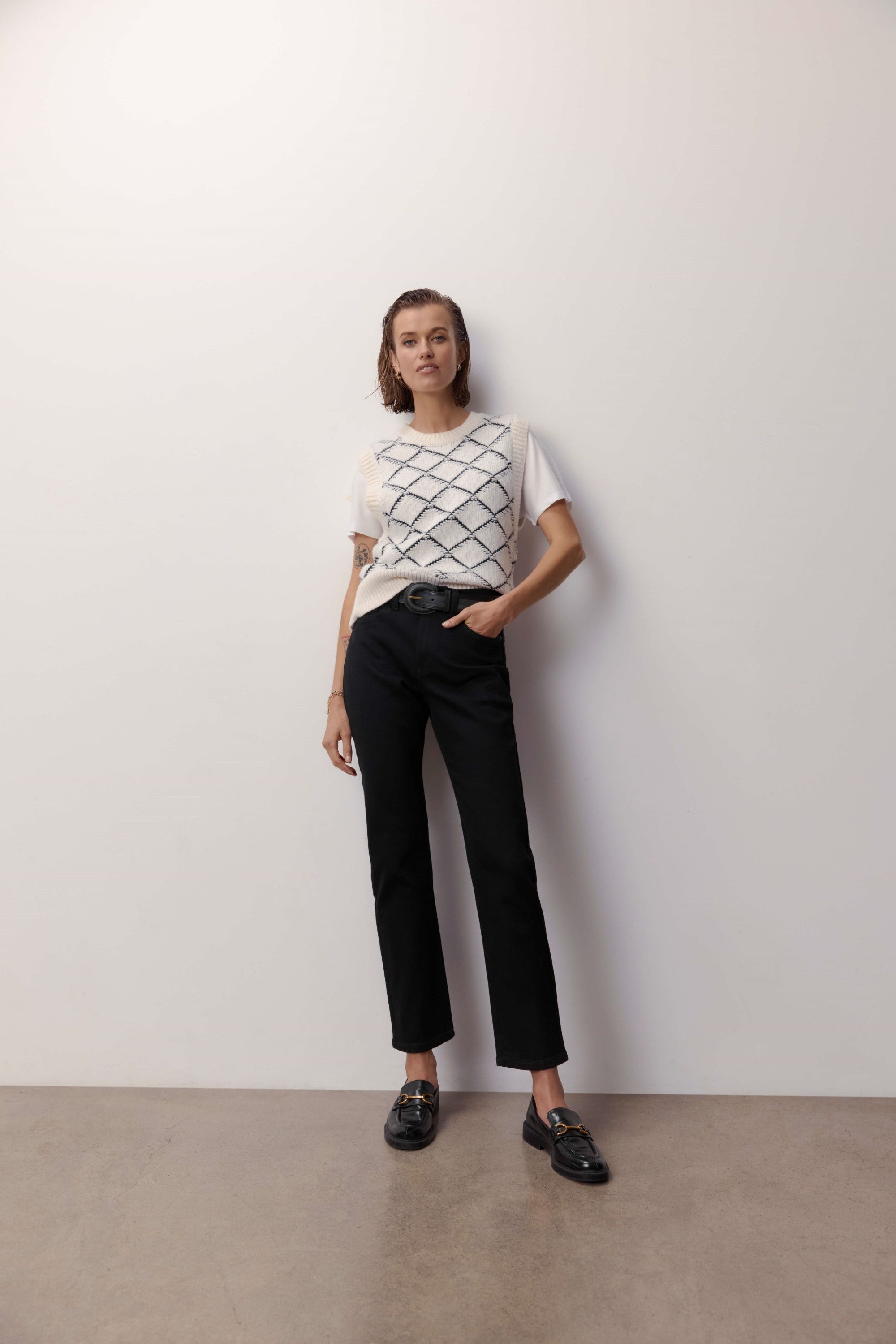 A person with short hair stands confidently against a plain white wall, wearing a white short-sleeve top with a black grid pattern, Max Georgia Slim Jeans in sustainable denim, and black loafers.