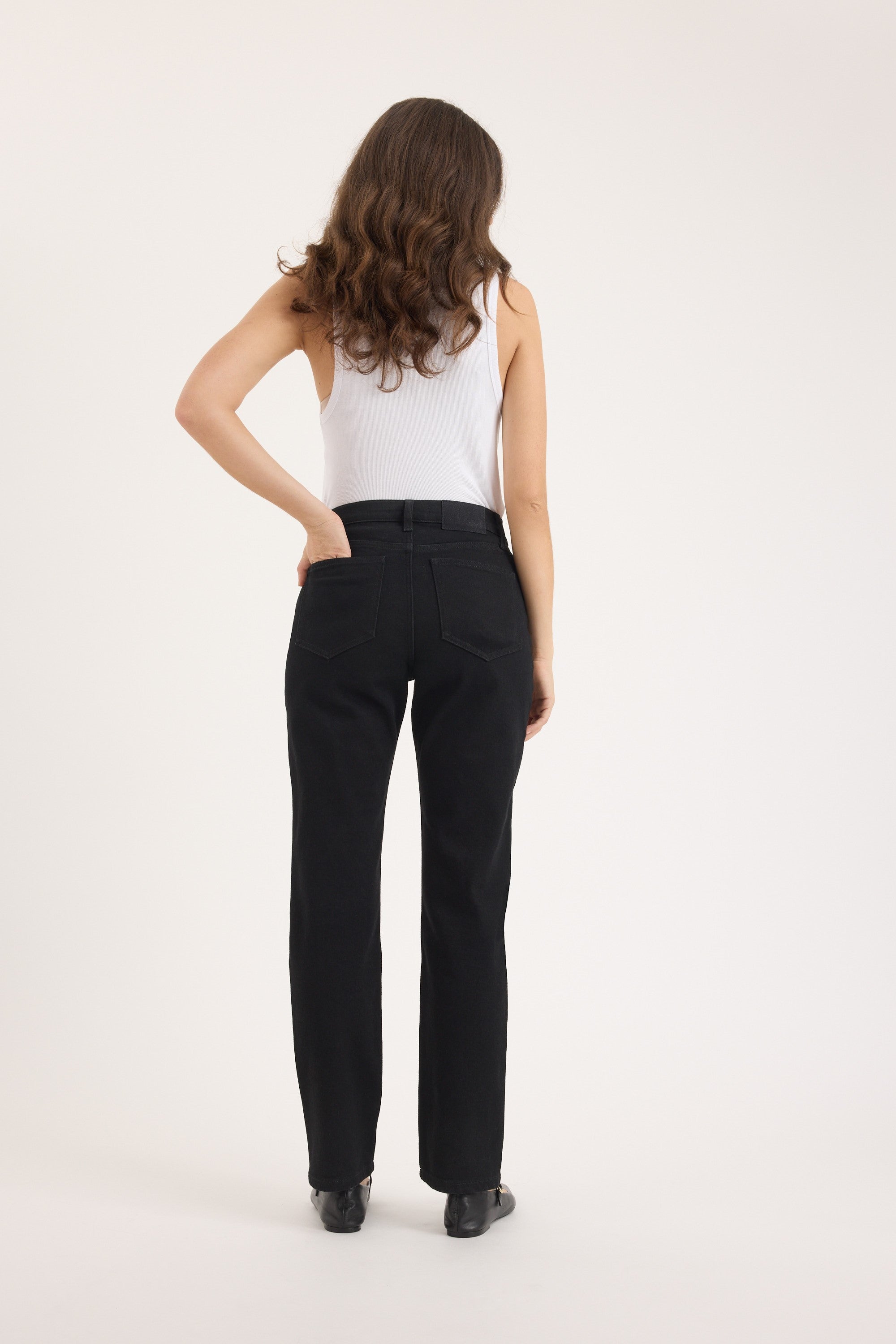 A person with long wavy brown hair, wearing a white sleeveless top and Max Georgia Slim Jeans, stands facing away with one hand in a back pocket against a plain light background.