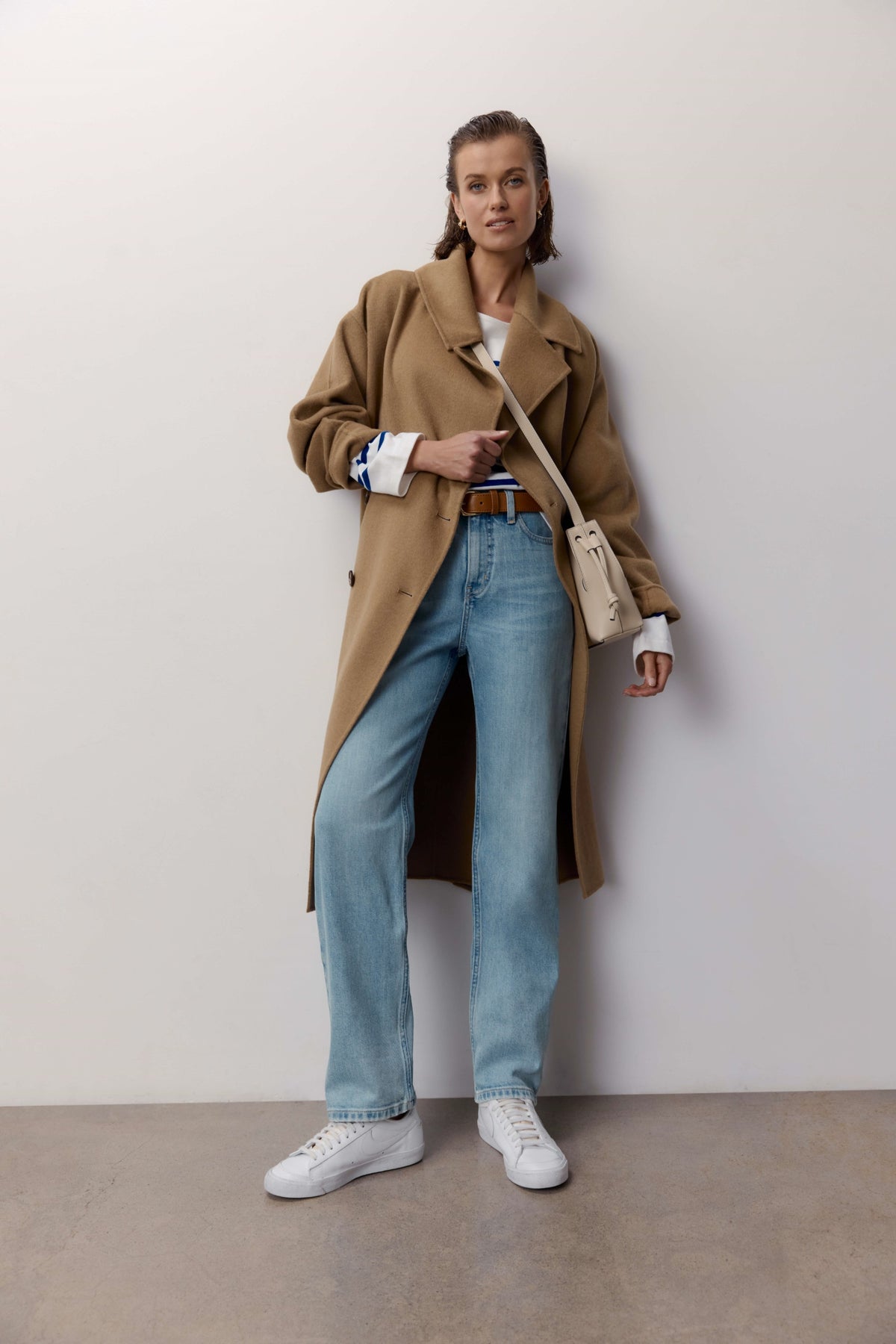 Woman in Arizona Straight Jeans by Max, tan coat, and white sneakers stands by a plain wall.