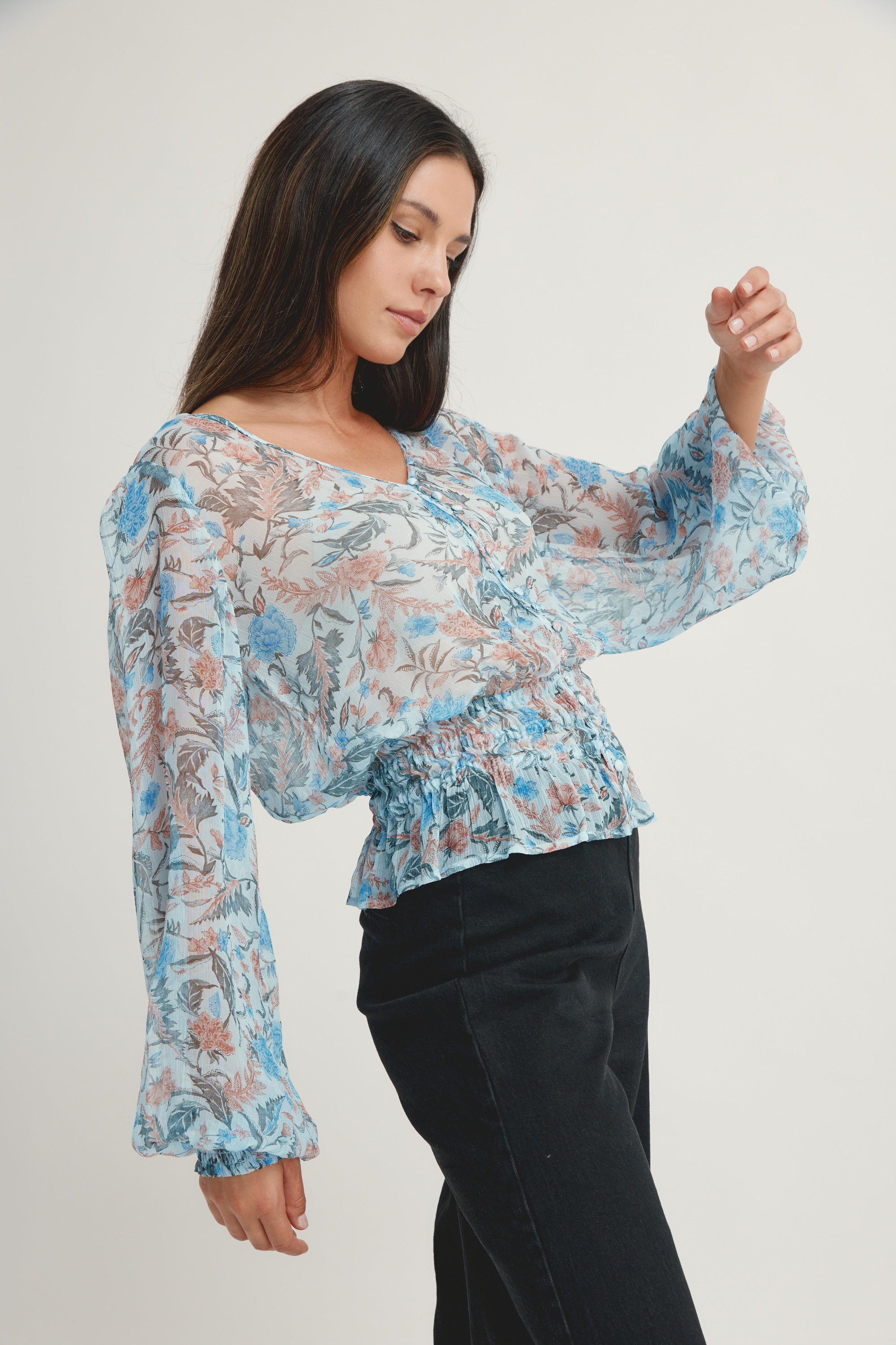A woman with long dark hair models the Max Joan Button Front Blouse, featuring a sheer floral print in blue and brown, paired with black pants, as she stands in a relaxed pose against a plain white background.