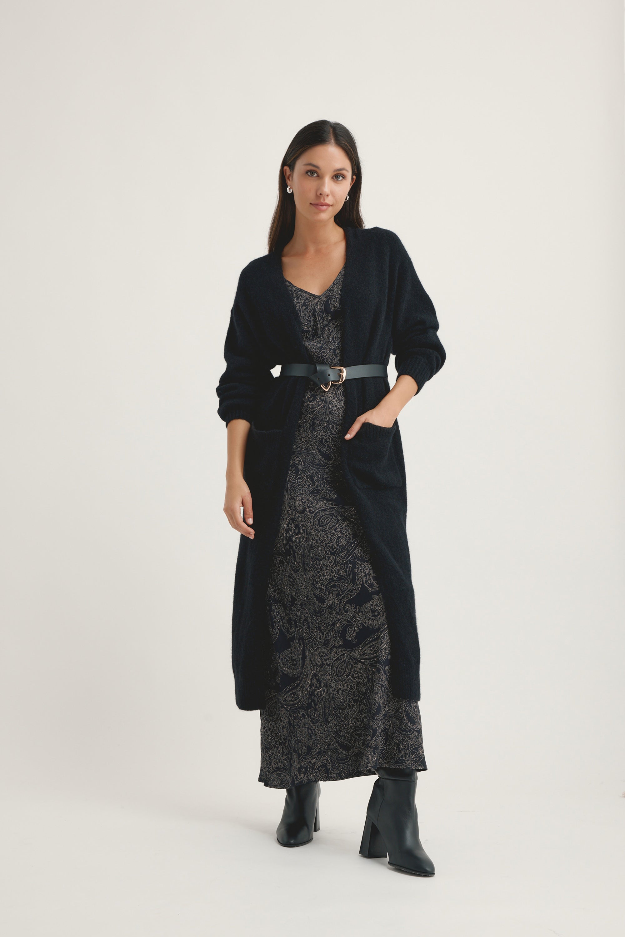 A woman with long dark hair and a neutral expression stands against a plain background wearing Max’s Fiona Slip Dress in paisley, paired with a dark belted open-front cardigan, black heeled boots, and hoop earrings.