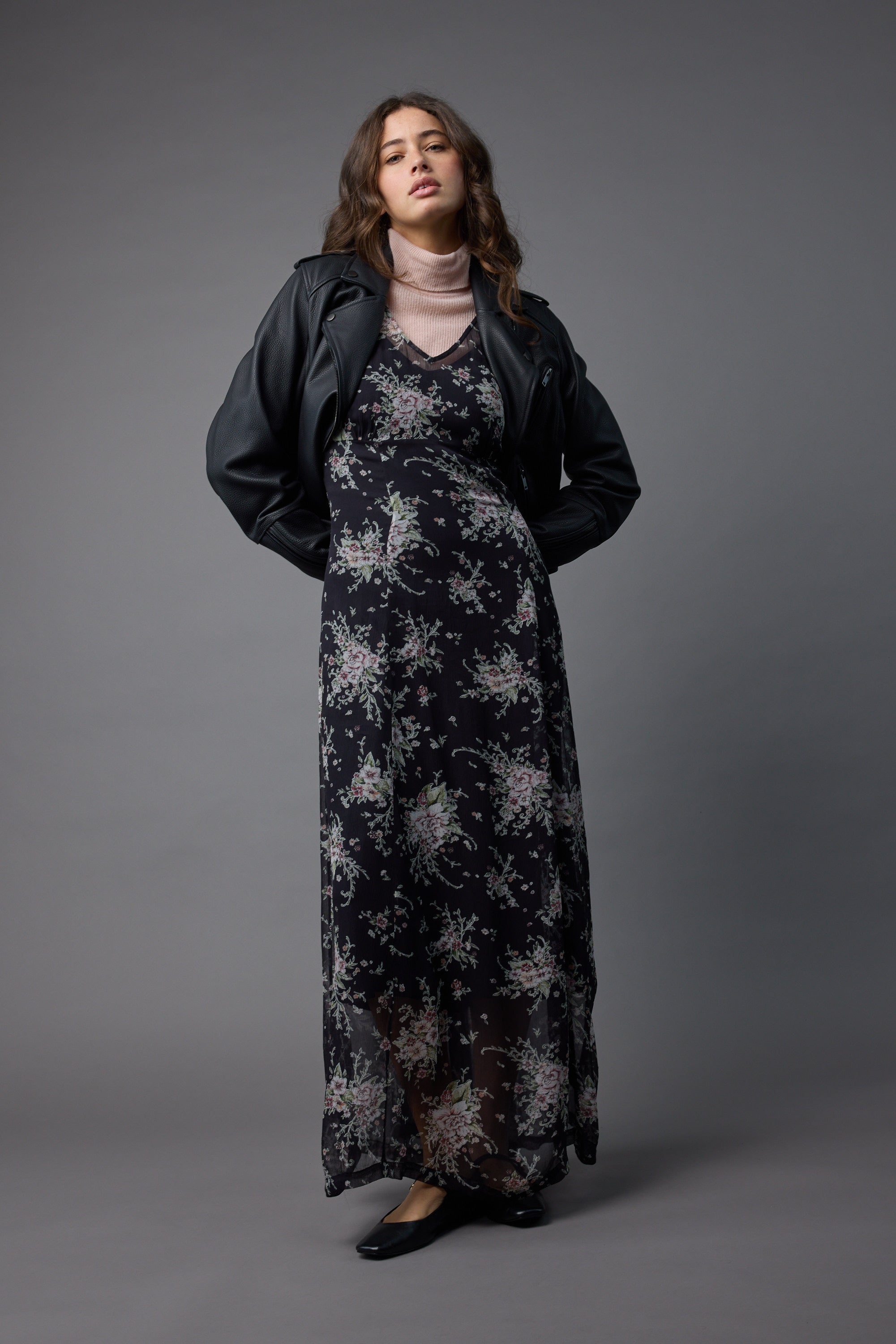 A woman poses against a gray background in the Max Amber Long Sleeve Dress, styled over a light pink turtleneck with a black leather jacket and flats. She stands with hands behind her back, gazing slightly upward.