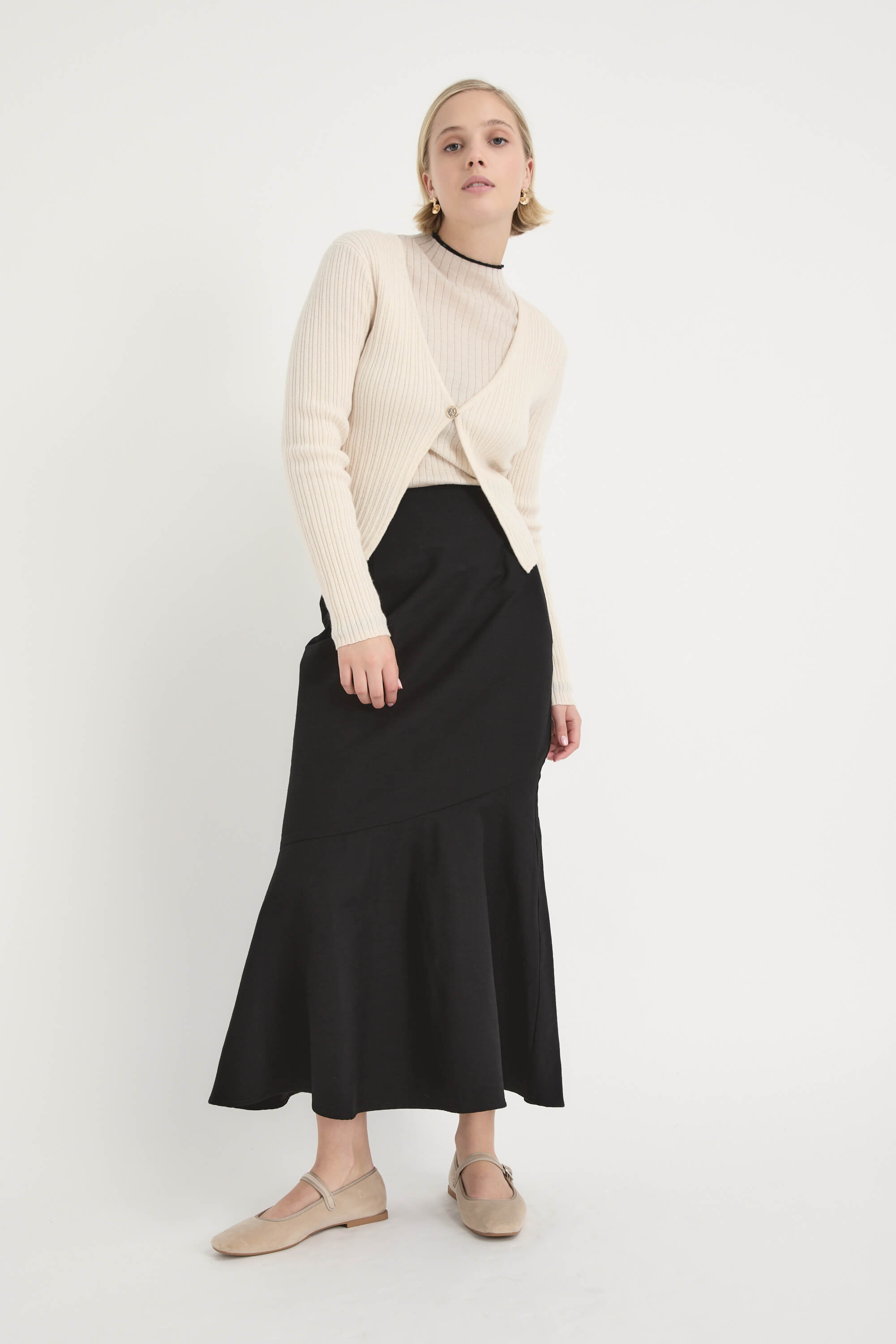 A woman with short blonde hair stands against a white background, wearing the Max Frances Flared Skirt paired with a cream ribbed cardigan, beige top, and beige shoes. She looks slightly upward in this elegant midi-length outfit.