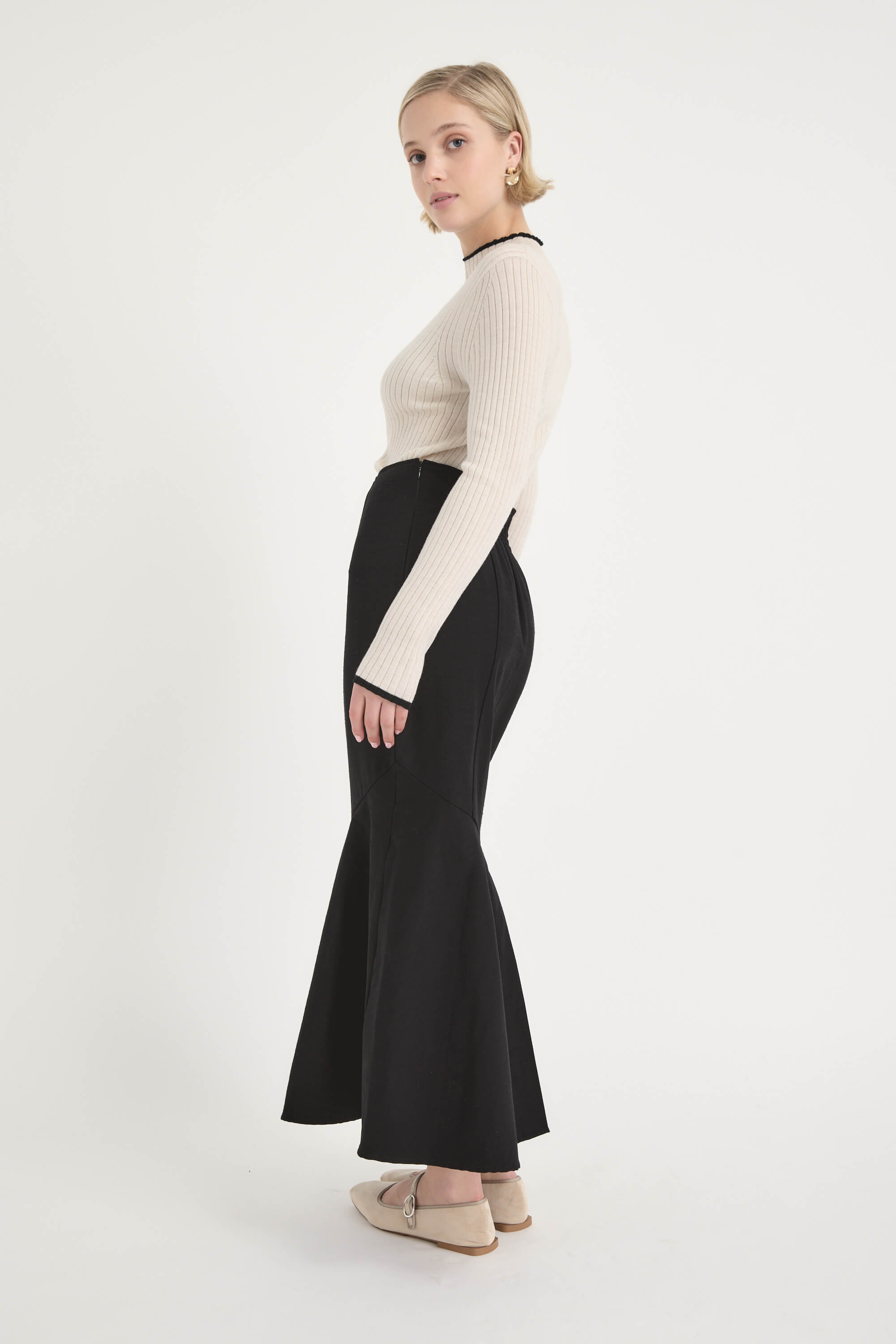 A woman in profile against a white background wears a fitted cream ribbed sweater, the Frances Flared Skirt by Max—a high-waisted black midi skirt—and beige heels. She accessorizes with gold hoop earrings and has short blonde hair.