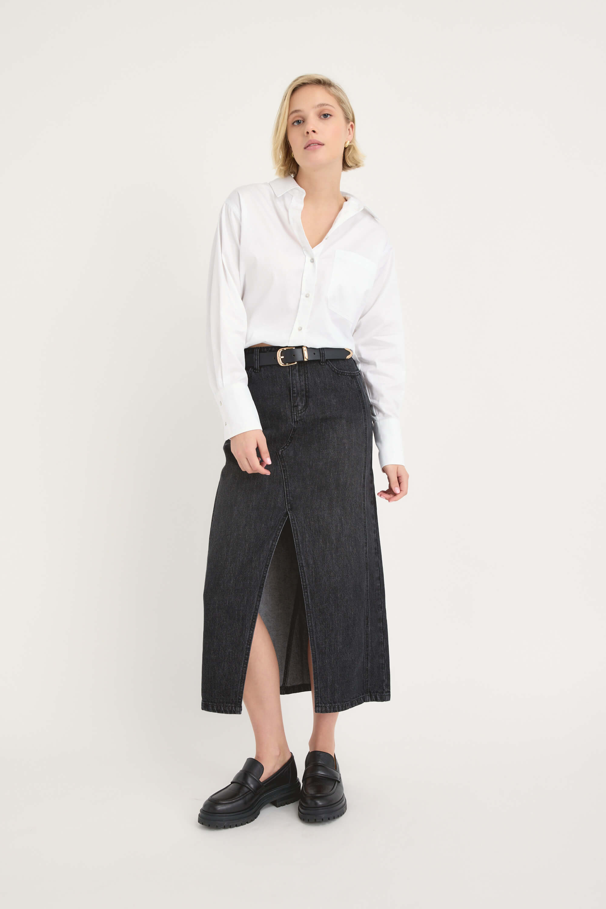 A person wearing a white button-up shirt, the Max Paris Maxi Denim Skirt with a front slit and black belt, and black loafers stands against a plain white background.