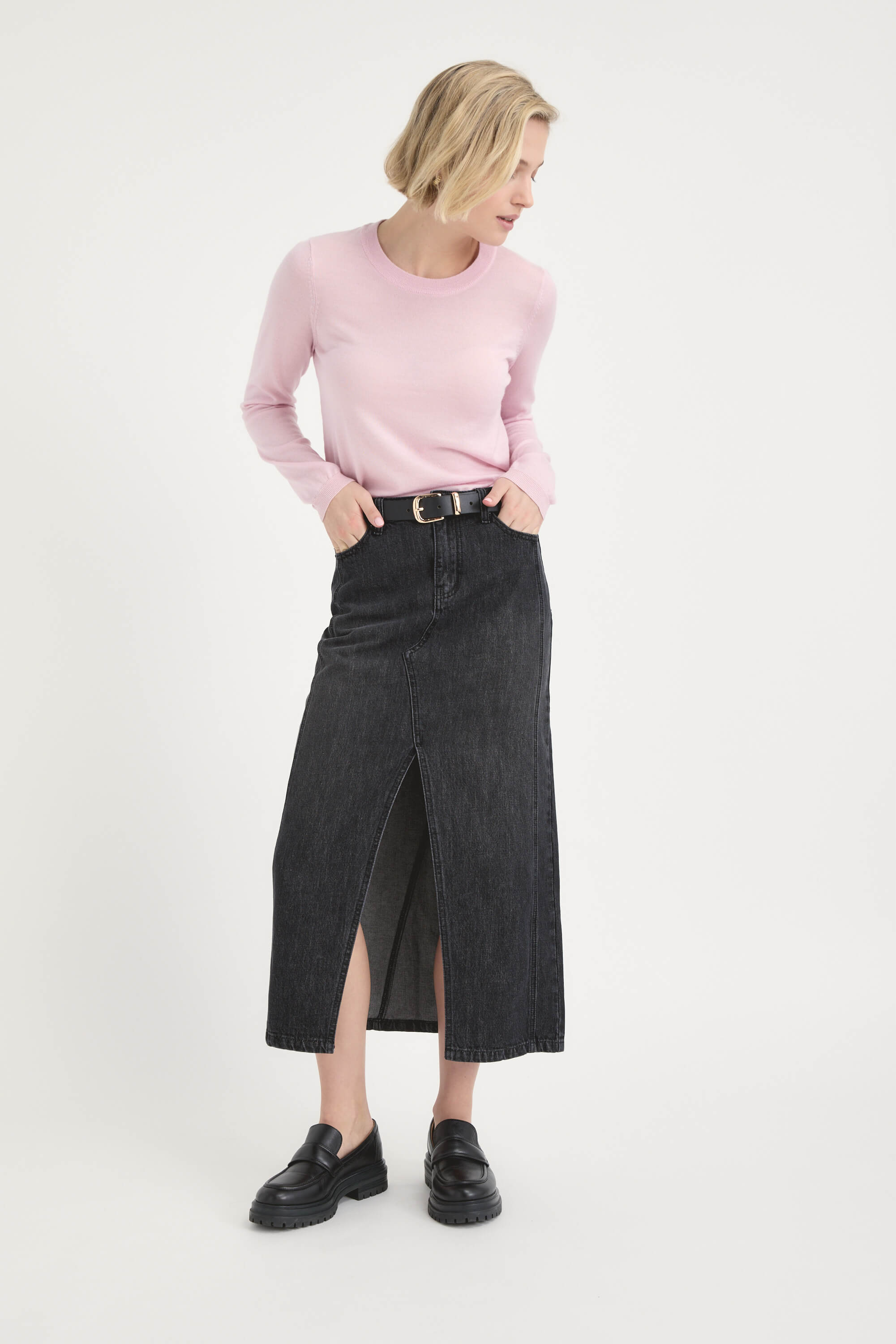 A woman in a light pink long-sleeve shirt, black loafers, and the Max Paris Maxi Denim Skirt with a front slit and belt stands against a plain white background, gazing slightly to her left.