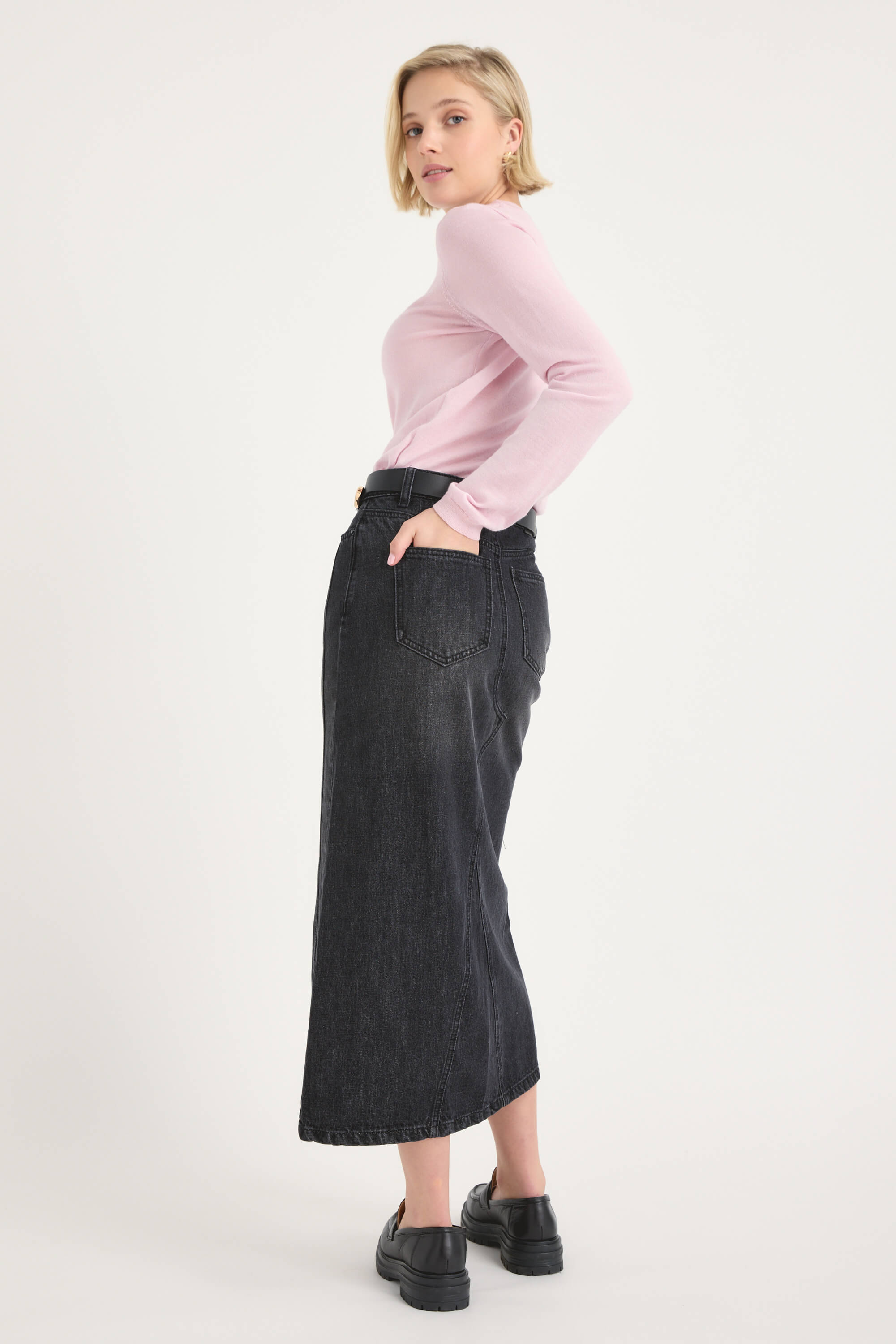 A person stands in profile against a white background, wearing a light pink long-sleeve top, black shoes, and the Max Paris Maxi Denim Skirt.