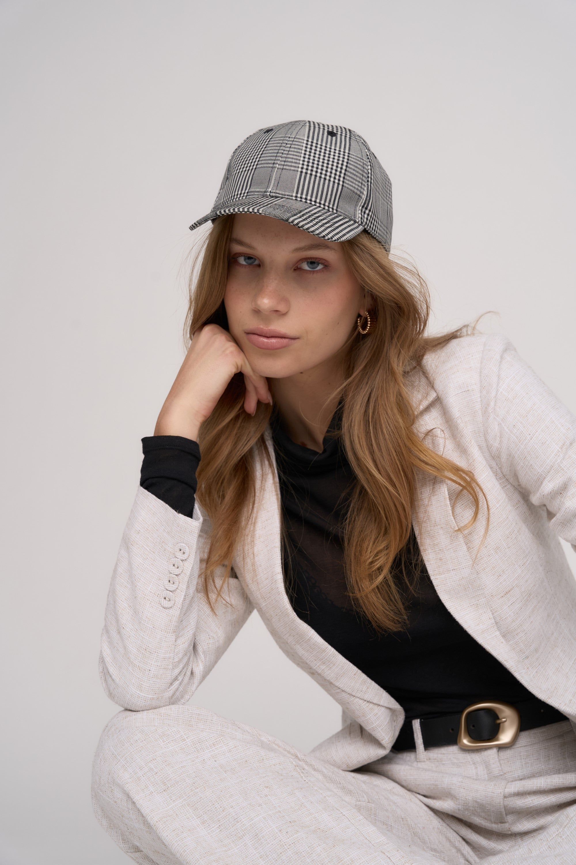 A woman with long blonde hair wears the Max Cherie Check Cap—a stylish wool blend cap for sun protection—paired with a light suit, black top, and gold hoops as she sits confidently against a plain background.