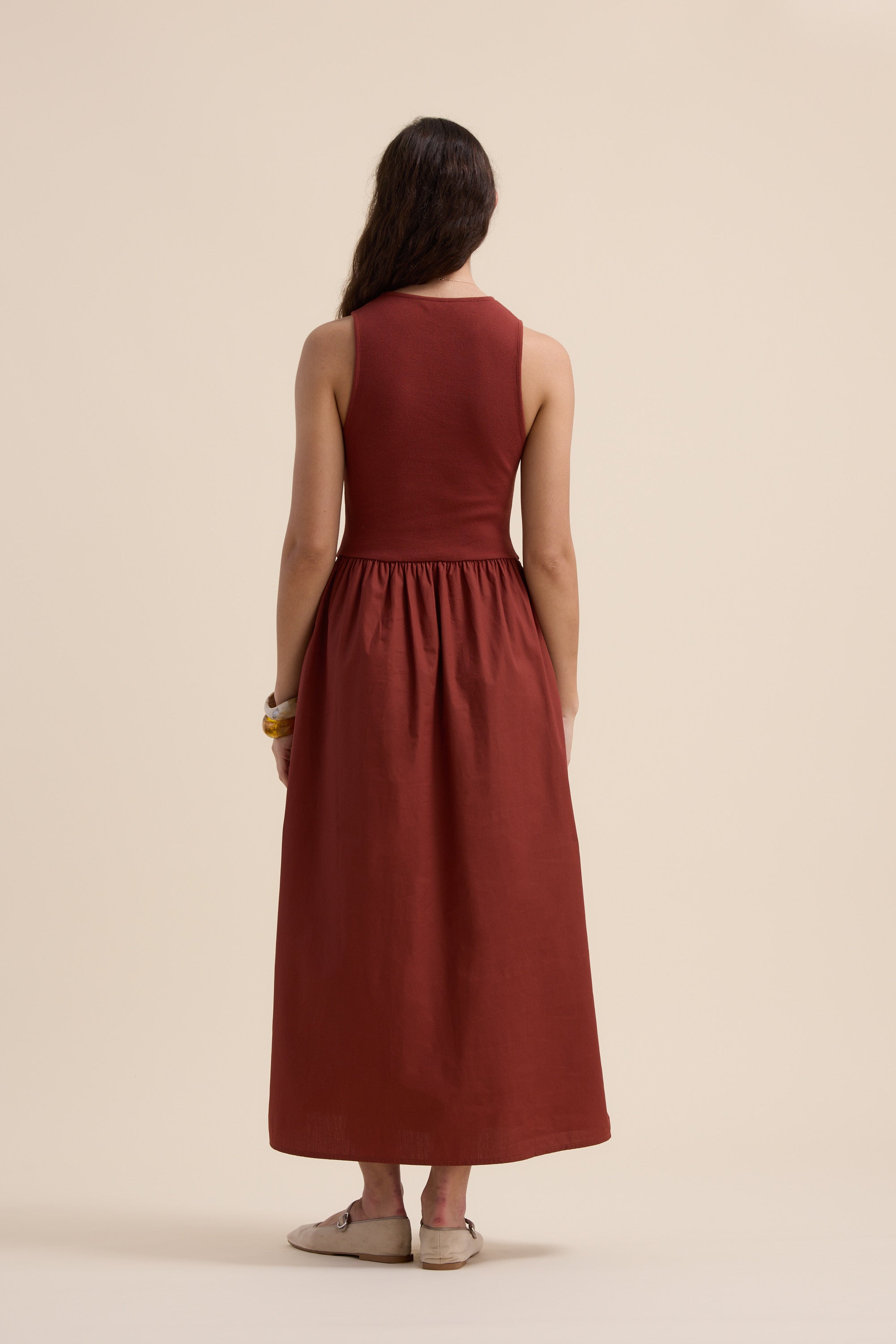 A woman with long dark hair wears Max's The One And Done Dress, a rust-colored organic cotton maxi with a fitted top and gathered skirt—an elegant, versatile staple. She stands against a beige background, accessorized with light shoes and bracelets.
