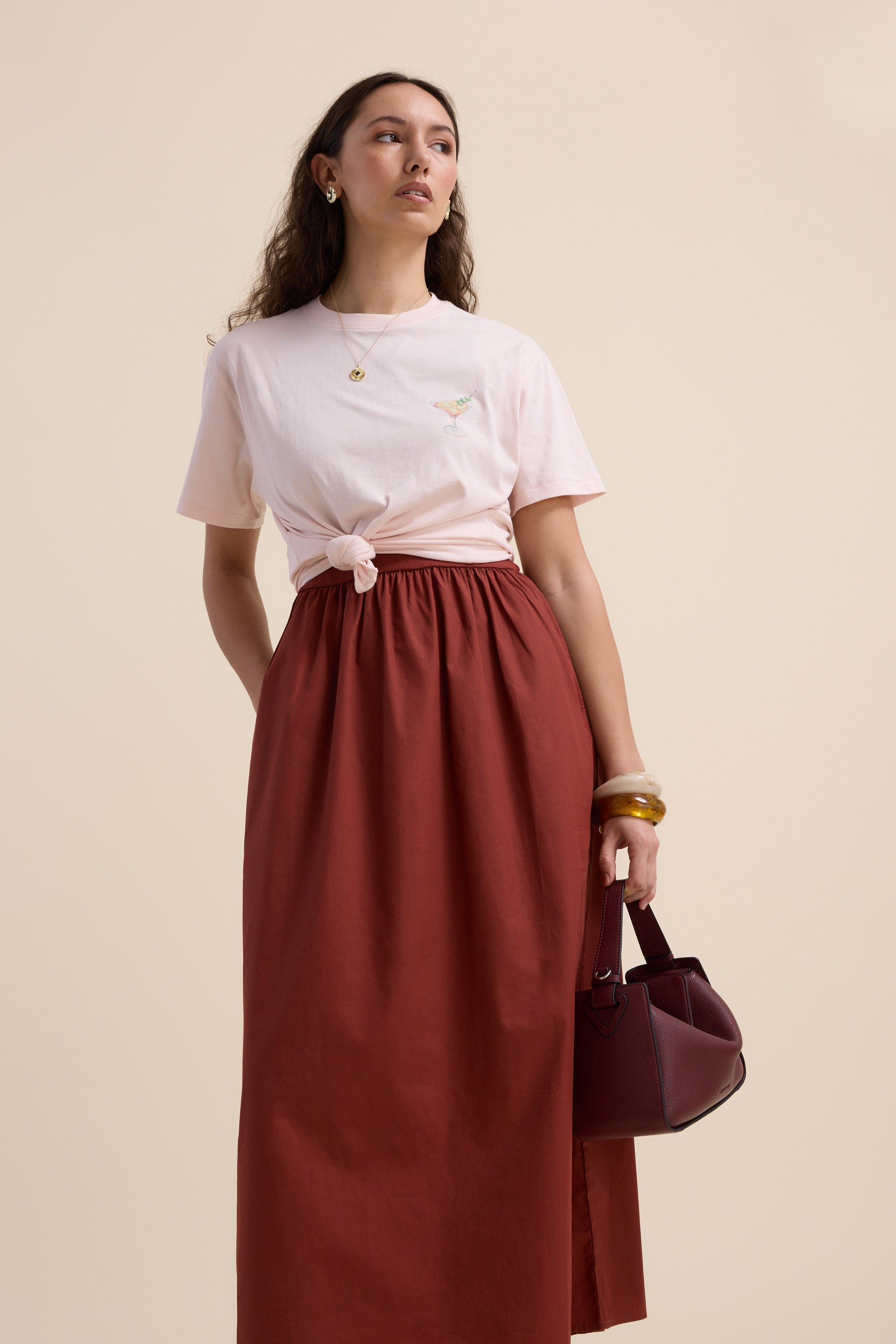 A woman poses against a beige background, wearing The One And Done Dress by Max, styled with gold jewelry, a chunky bangle, and holding a burgundy handbag by her side.
