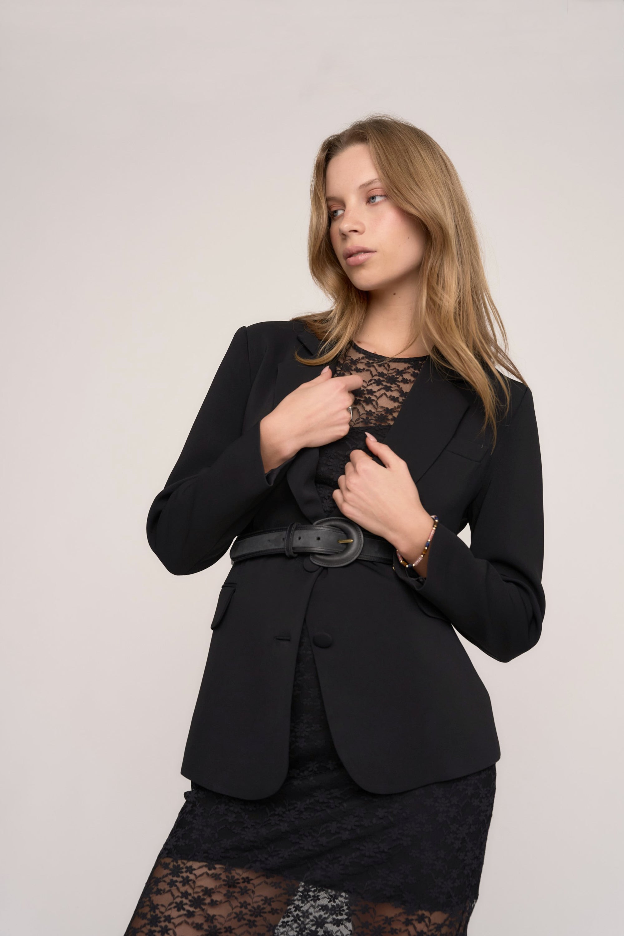 A woman with long, light brown hair models the Max Morgan Lace Dress, styled under a black belted blazer, standing against a plain light background and gazing slightly to the side.