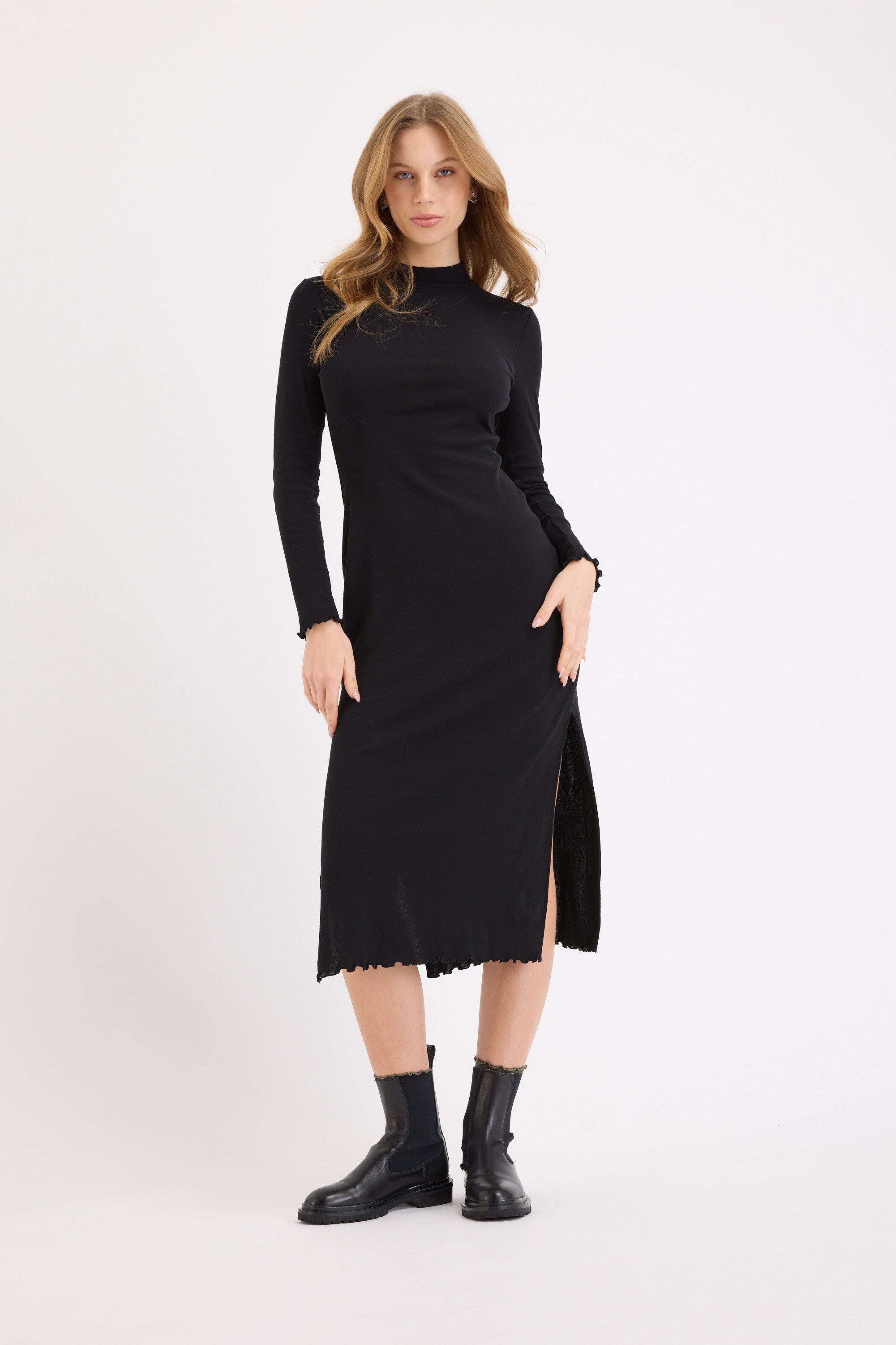 A woman poses against a plain white backdrop in the Max Nichole High Neck Layering Dress—a black, long-sleeved midi with side slit—paired with black ankle boots. Her hair is loose, and she places a hand on her hip for a chic winter look.