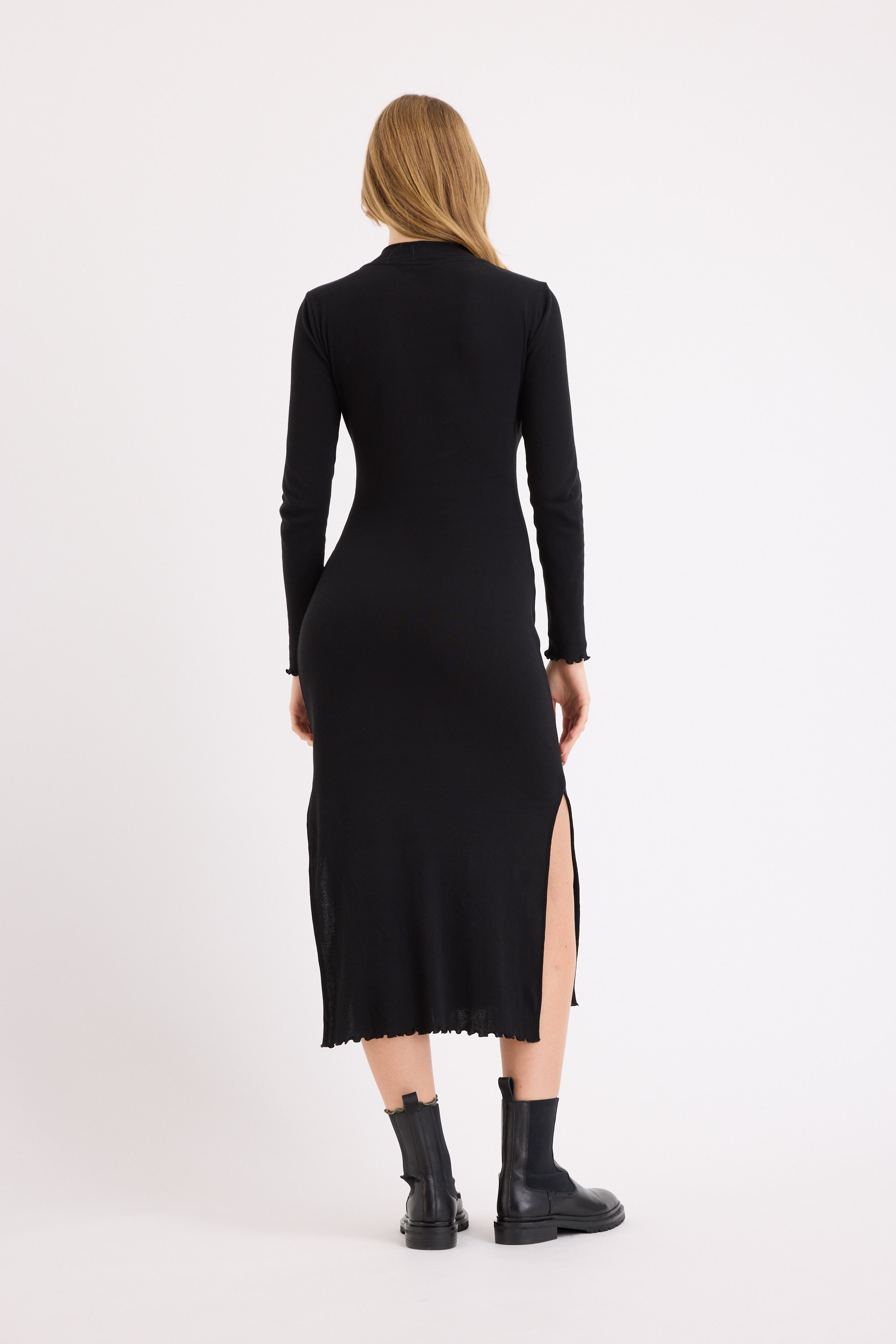 A woman faces away, wearing the Max Nichole High Neck Layering Dress—an ankle-length pima cotton piece with long sleeves and side slits—paired with black ankle boots. Her blonde hair is down, completing a chic winter look against a white background.
