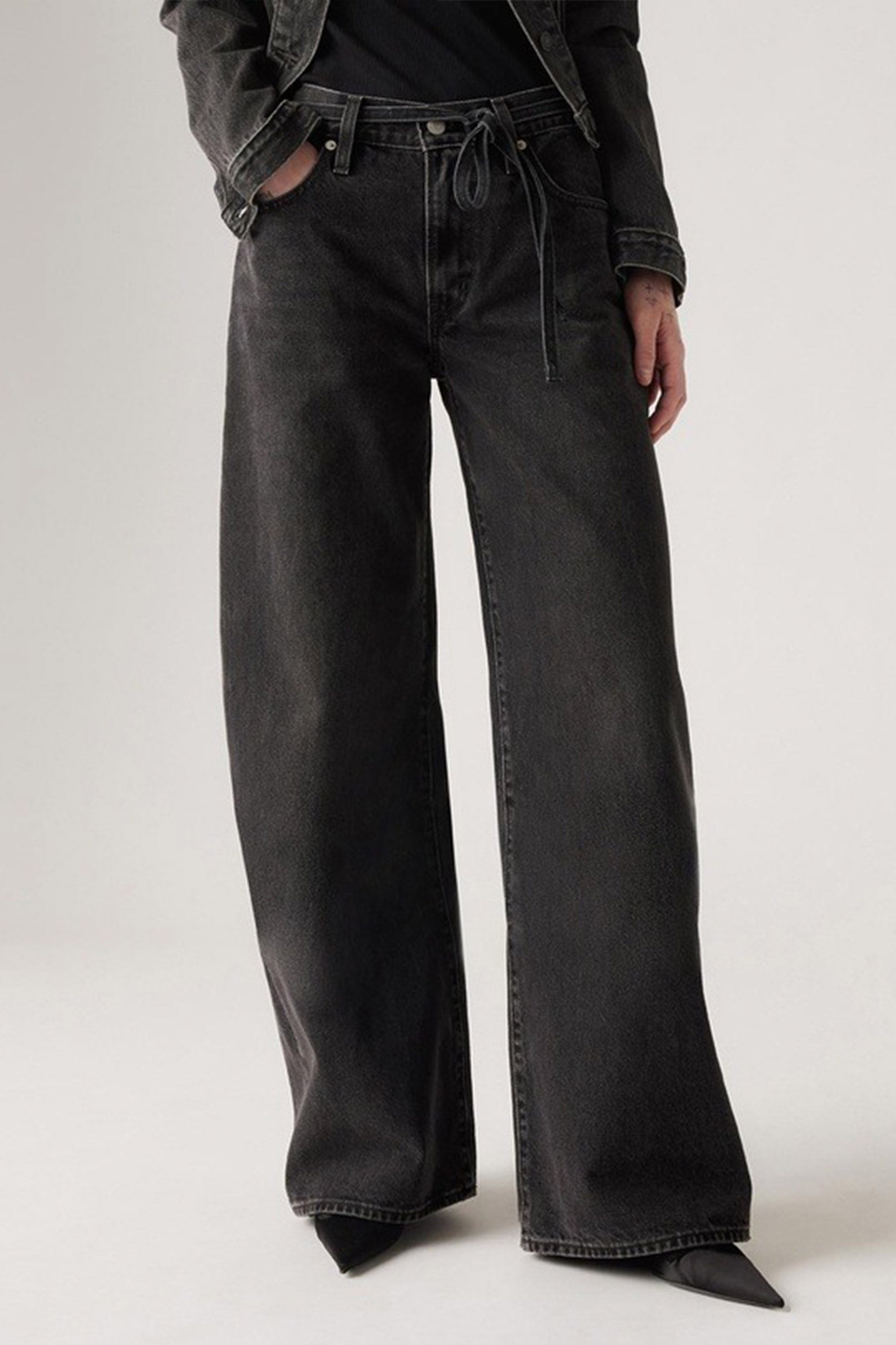 A person models Levi's XL Straight Jeans by Levi's—high-waisted, wide-leg black denim with an adjustable tie waistband—paired with pointed black shoes and a dark jacket, set against a neutral background.