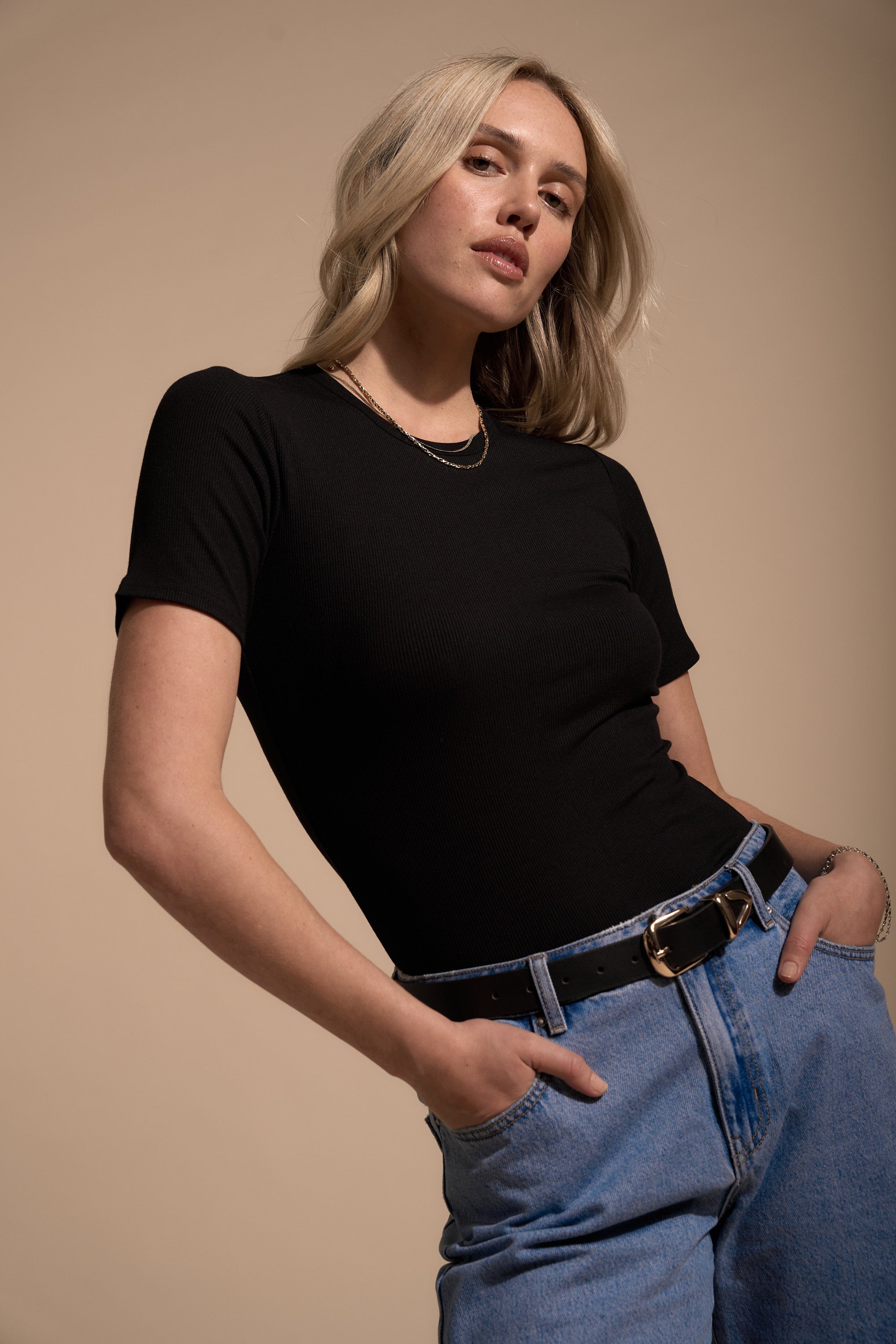 A person with shoulder-length blonde hair confidently models the Elementary Basics Rib Tee by Elementary, paired with blue jeans and a black belt, standing against a beige background to show how perfect this rib tee is for layering.