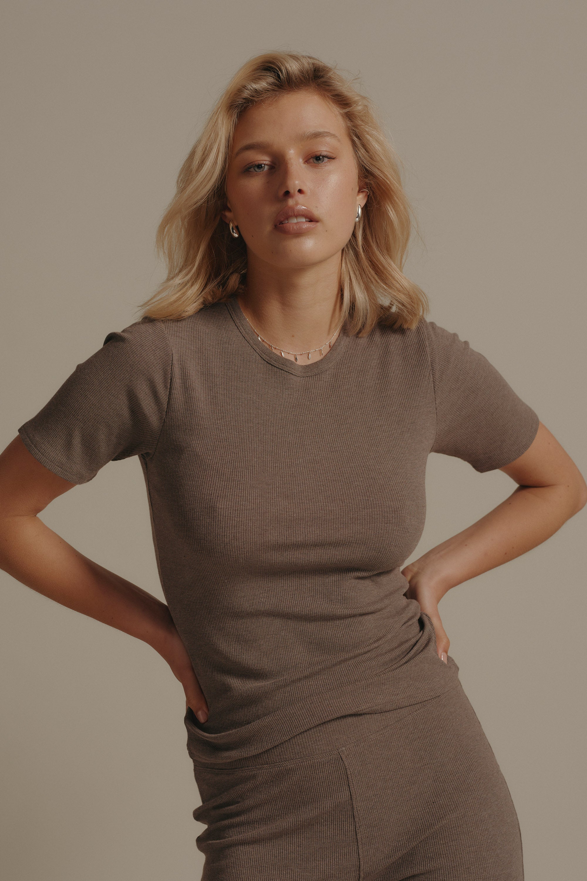 A blonde woman stands against a plain backdrop, confidently in the Elementary Basics Rib Tee by Elementary with matching pants—hands on hips, she epitomizes effortless style in this fitted short-sleeve essential.