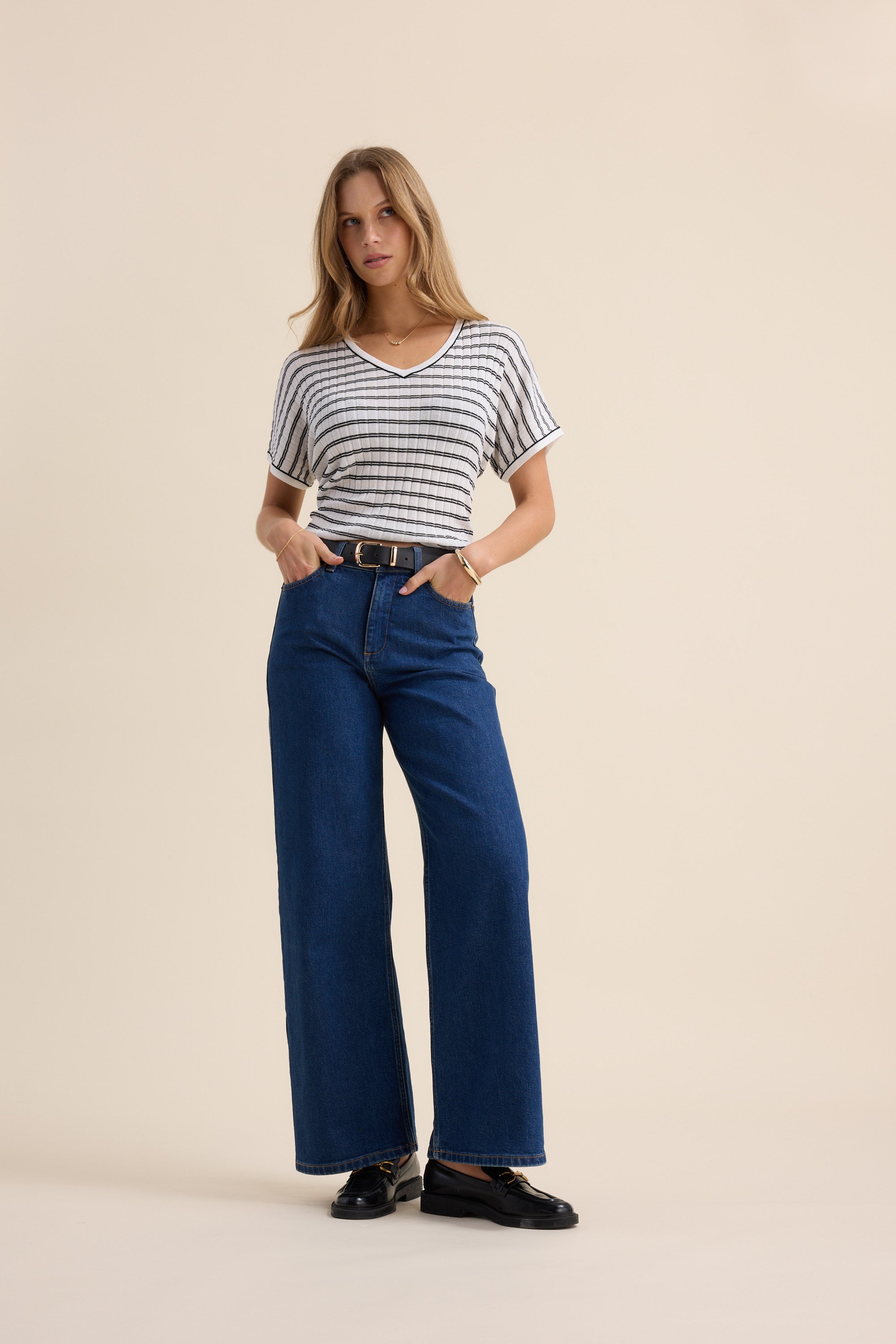 A woman stands against a plain background wearing The V Neck Knitted Top by Max, paired with high-waisted blue wide-leg jeans, a black belt, and black shoes. Her hands are in her pockets as she looks at the camera.