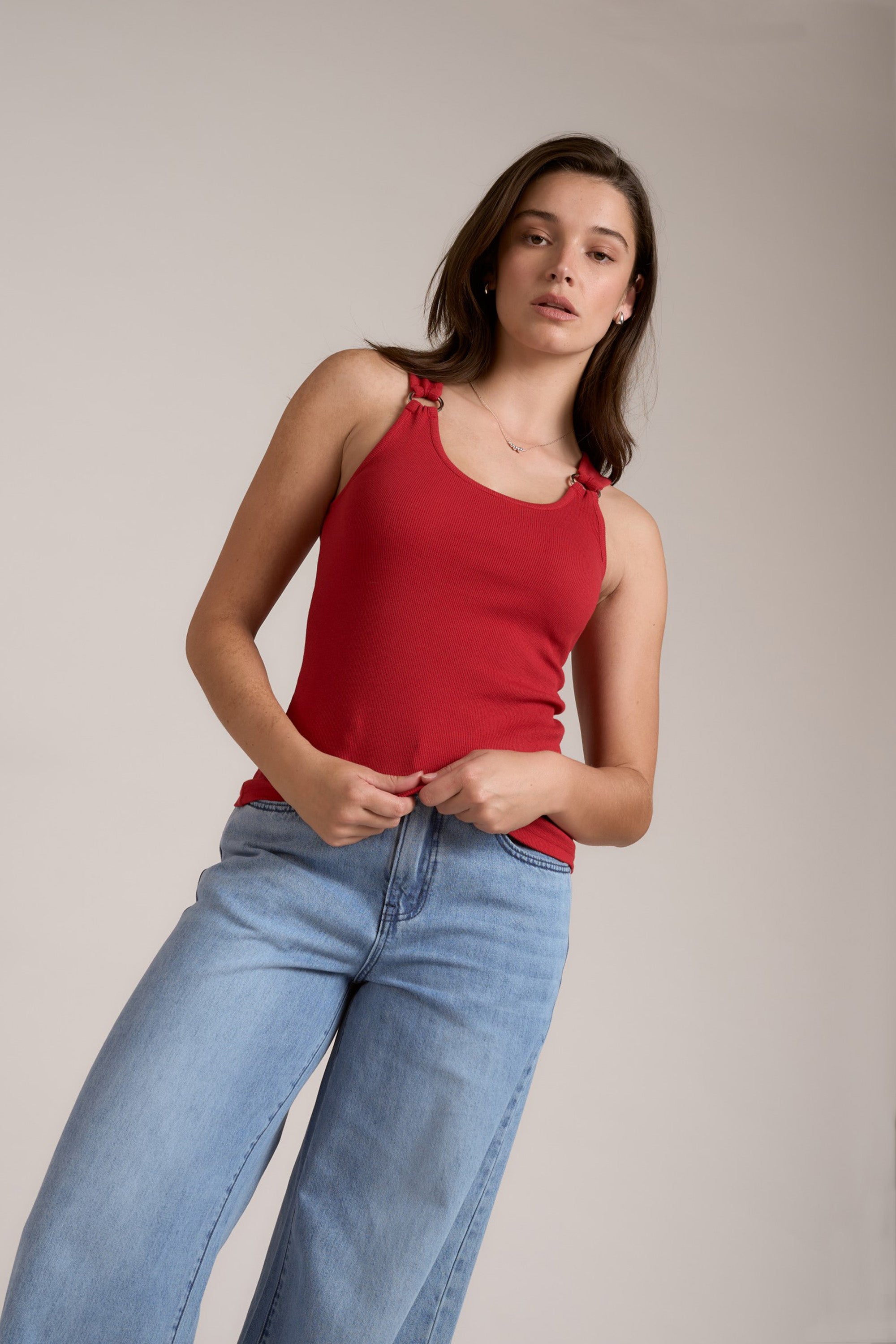 A young woman with straight brown hair wears the Max Eliza Ring Detail Tank, featuring silver hardware, and light blue jeans. She stands against a plain beige background, looking confidently at the camera.