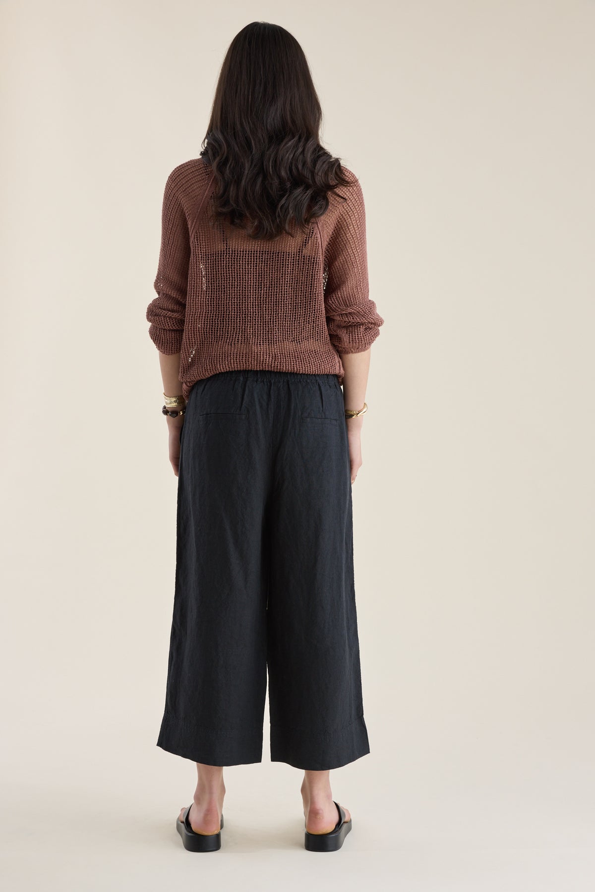 Person facing away in brown sweater, Elementary High Tide Linen Crop Pants, and sandals.