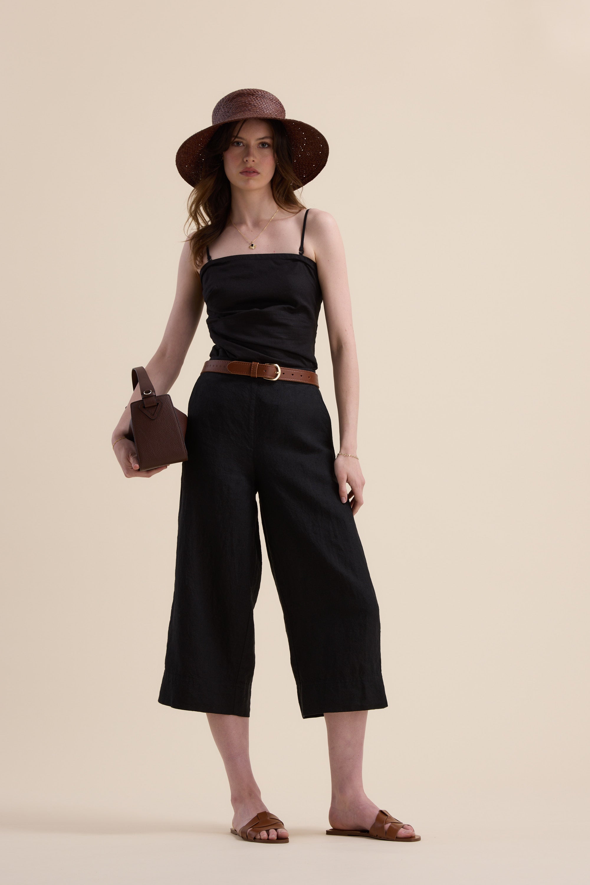 A woman stands against a beige background wearing Elementary’s High Tide Linen Crop, a black sleeveless top, wide-brimmed brown hat, brown belt, brown slide sandals, and holding a small brown bag.