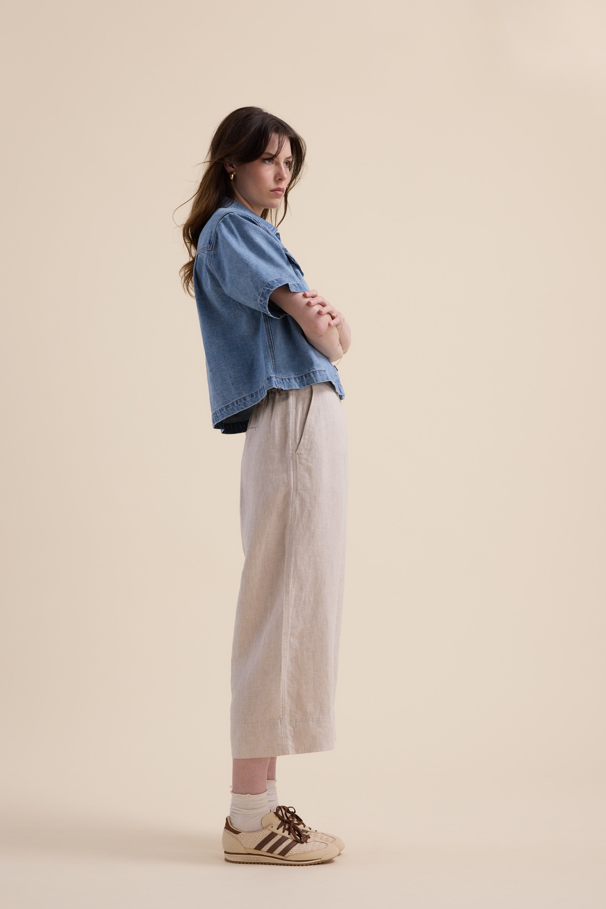 A woman stands in profile against a beige background, arms crossed, wearing Elementary’s High Tide Linen Crop pants with a blue denim shirt, white socks, and beige sneakers featuring brown stripes.