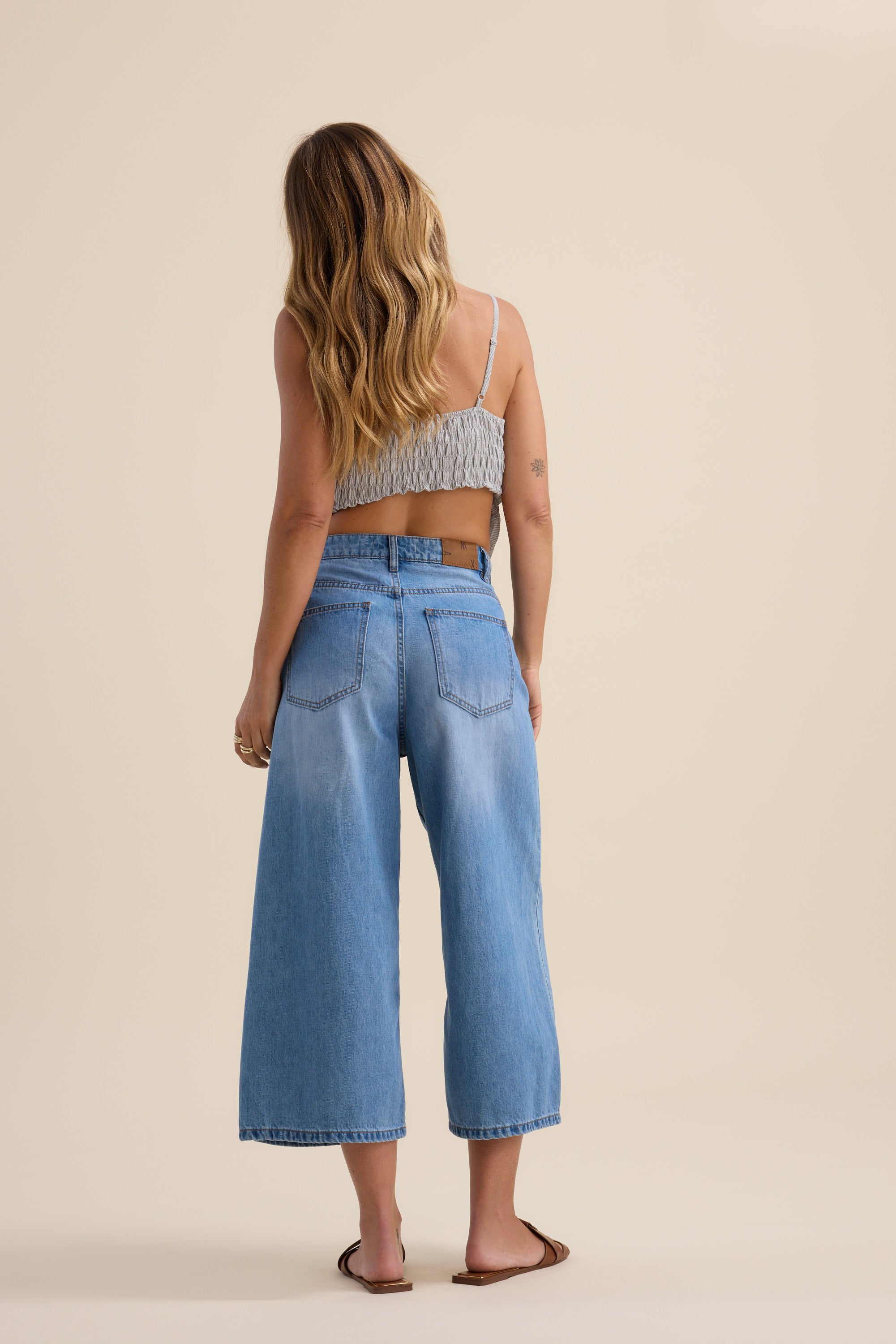 A woman with long wavy hair stands facing away, wearing a gray crop top, Max Derby Wide Leg Cropped Jeans, and brown sandals against a plain beige background.