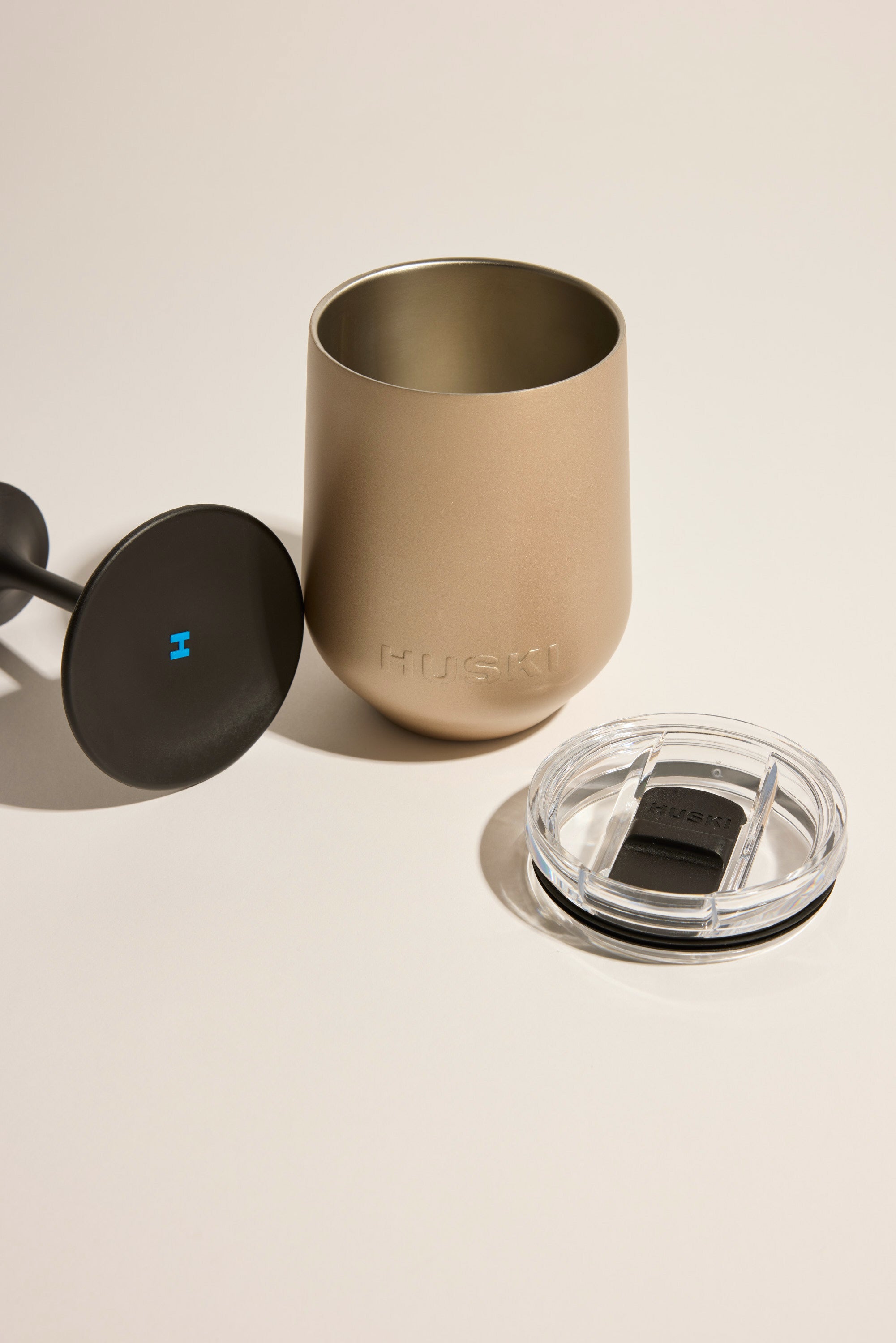 The matte-finish Huski Wine Tumbler 2.0 by Huski rests on a flat surface, with its black lid and transparent removable cover placed beside it; the tumbler features the embossed Huski brand name on its side.