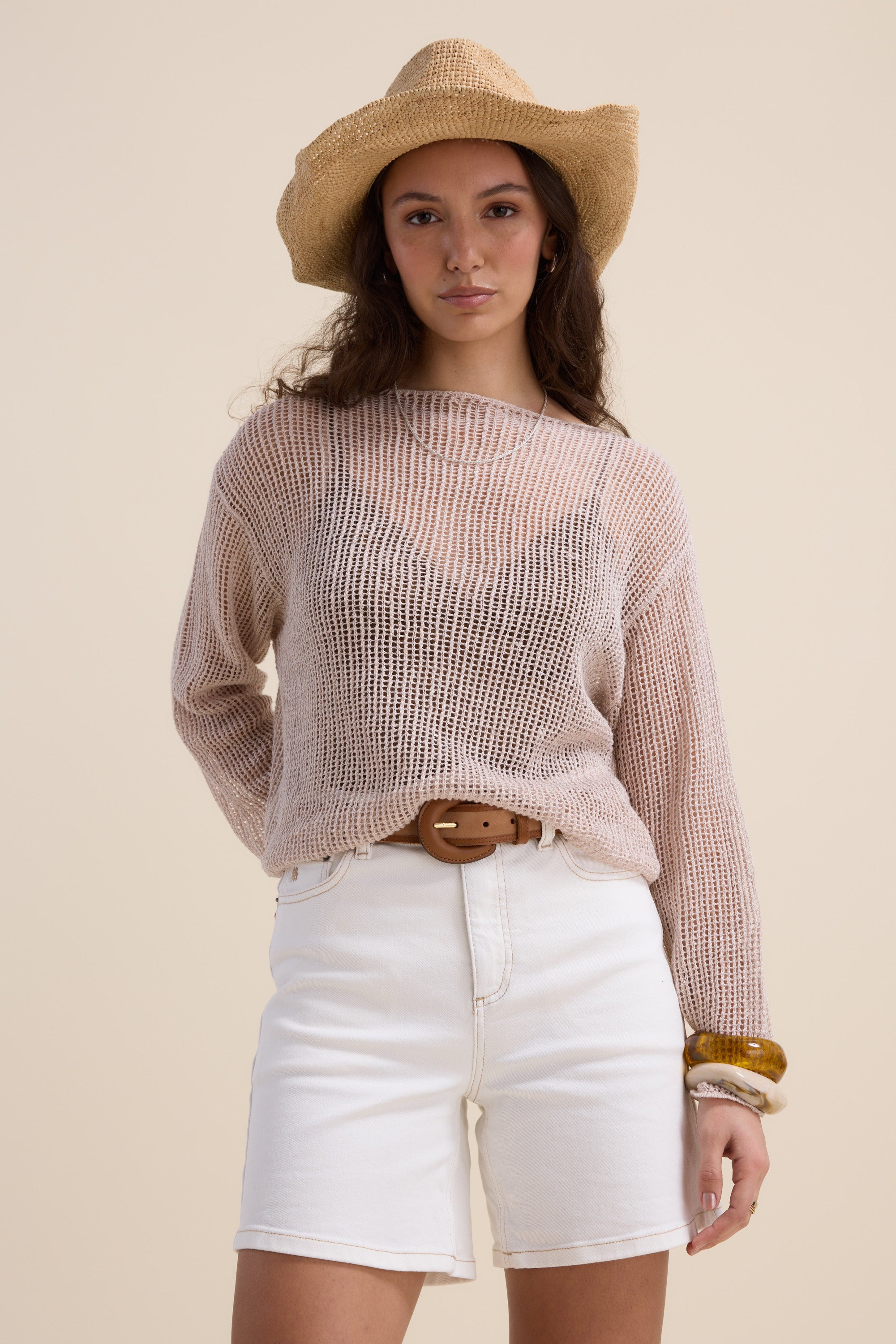 A woman wearing a straw hat, loose-knit beige sweater, and The Effortless Denim Short by Max with a brown belt stands against a plain background, one hand in her pocket, gazing confidently at the camera.