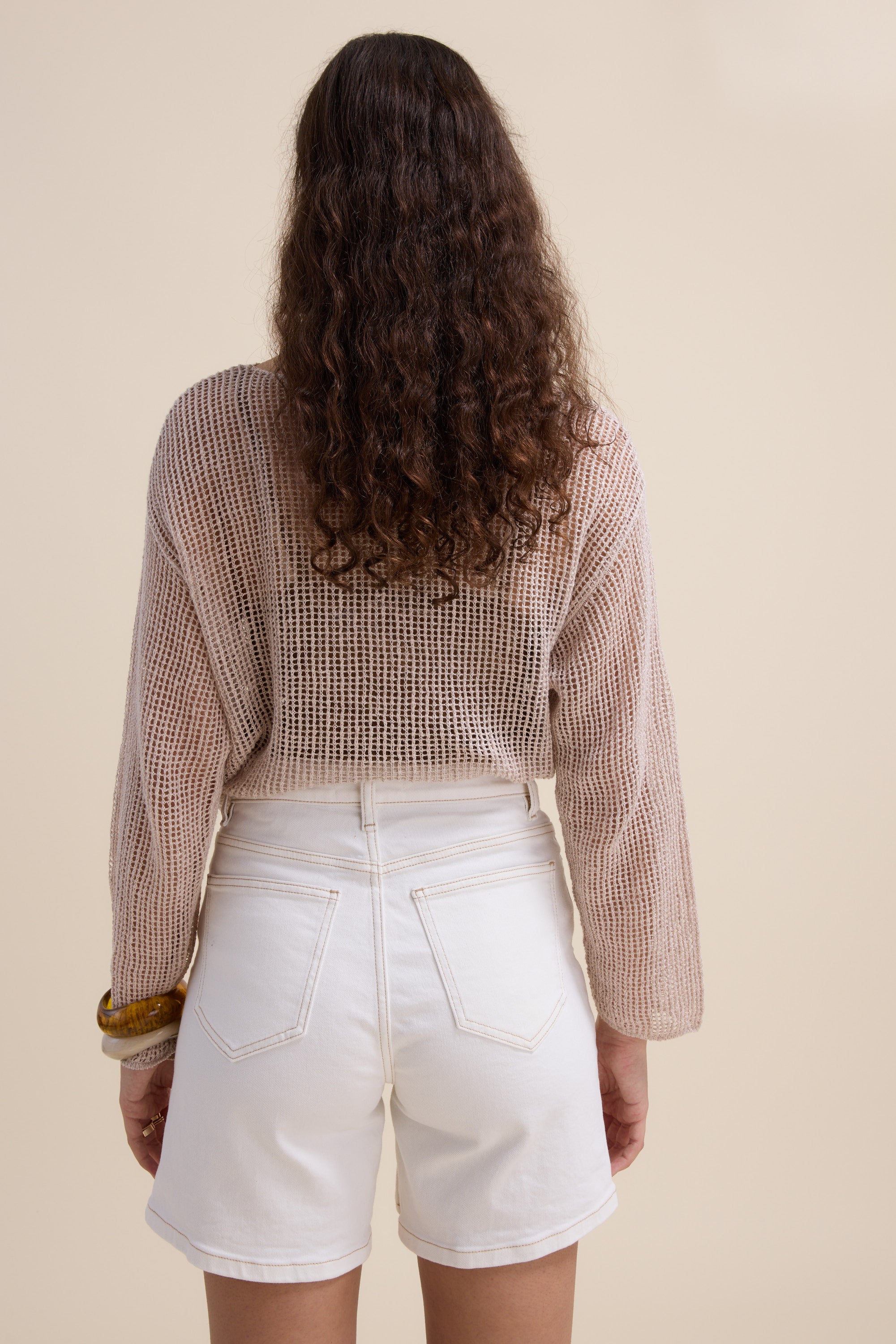 A woman with long curly brown hair, seen from behind, wears Max's The Effortless Denim Short with a loose beige knit sweater and a chunky brown bracelet. The background is plain beige.