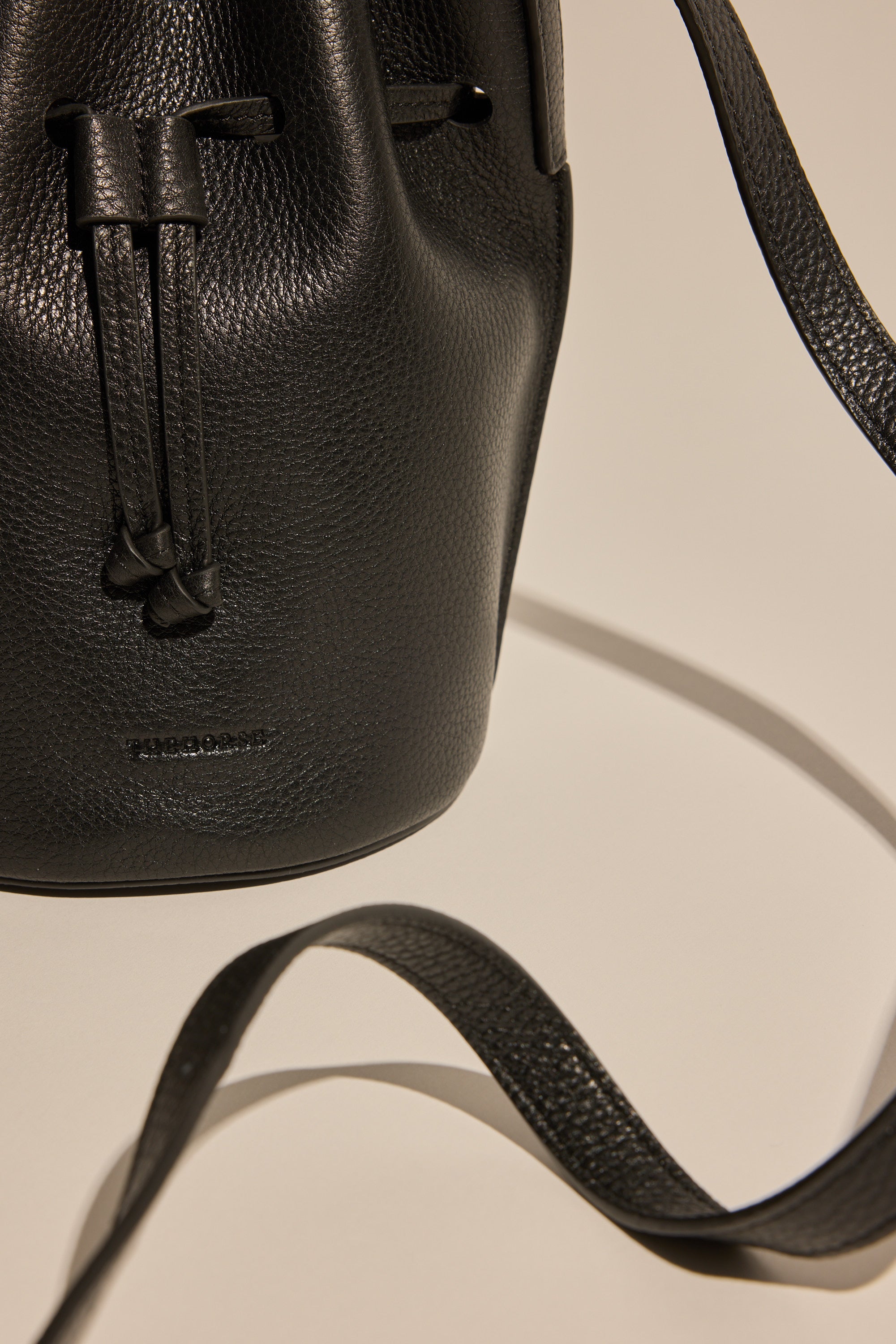 Close-up of The Horse Bob Bucket by The Horse, featuring black grained leather, a long curved crossbody strap, and textured surface, set against a neutral background.