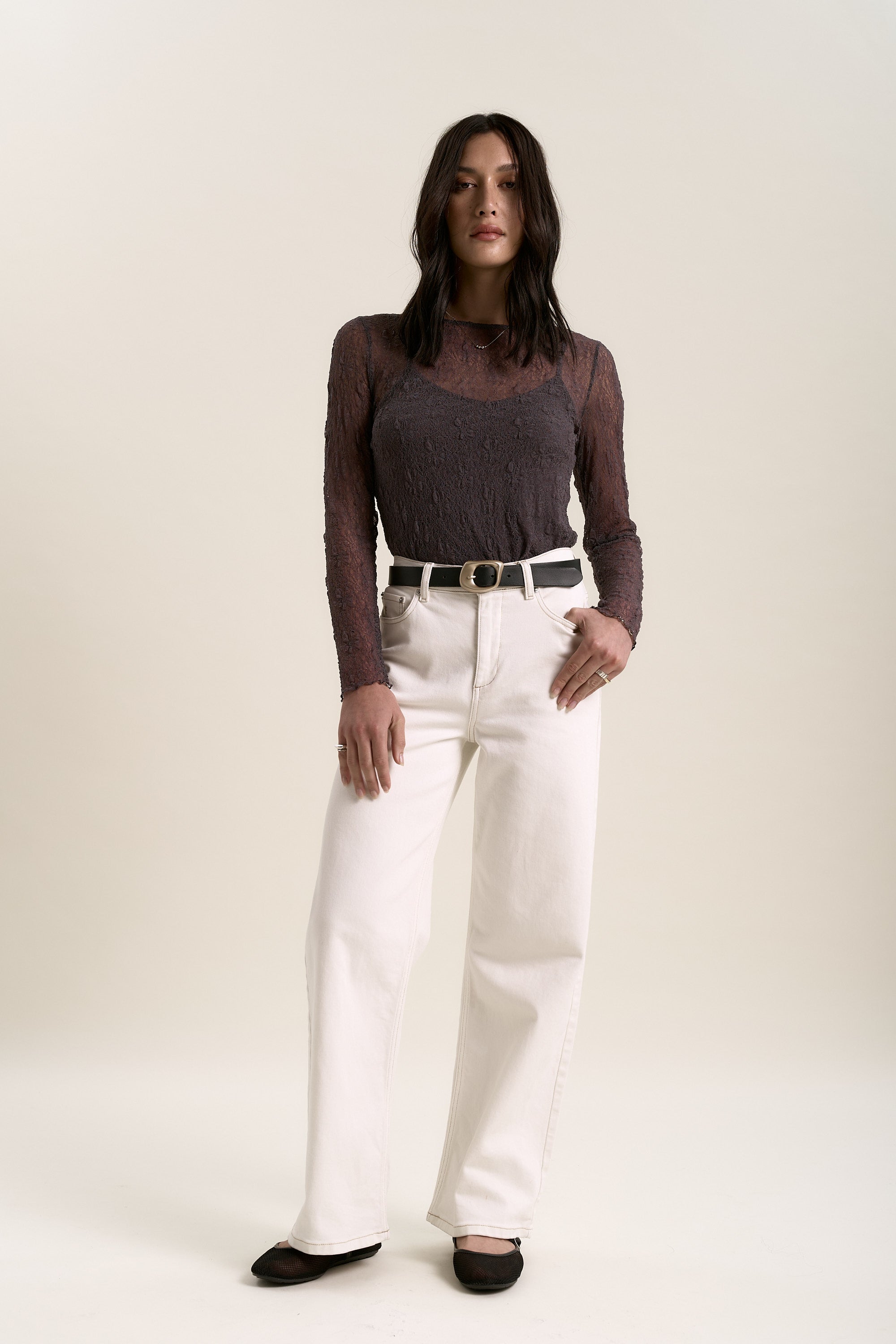 A woman with dark hair and a neutral expression stands against a plain background, wearing the Max Lavender Lace Top with removable cami, paired with white high-waisted pants, a black belt, and black shoes.