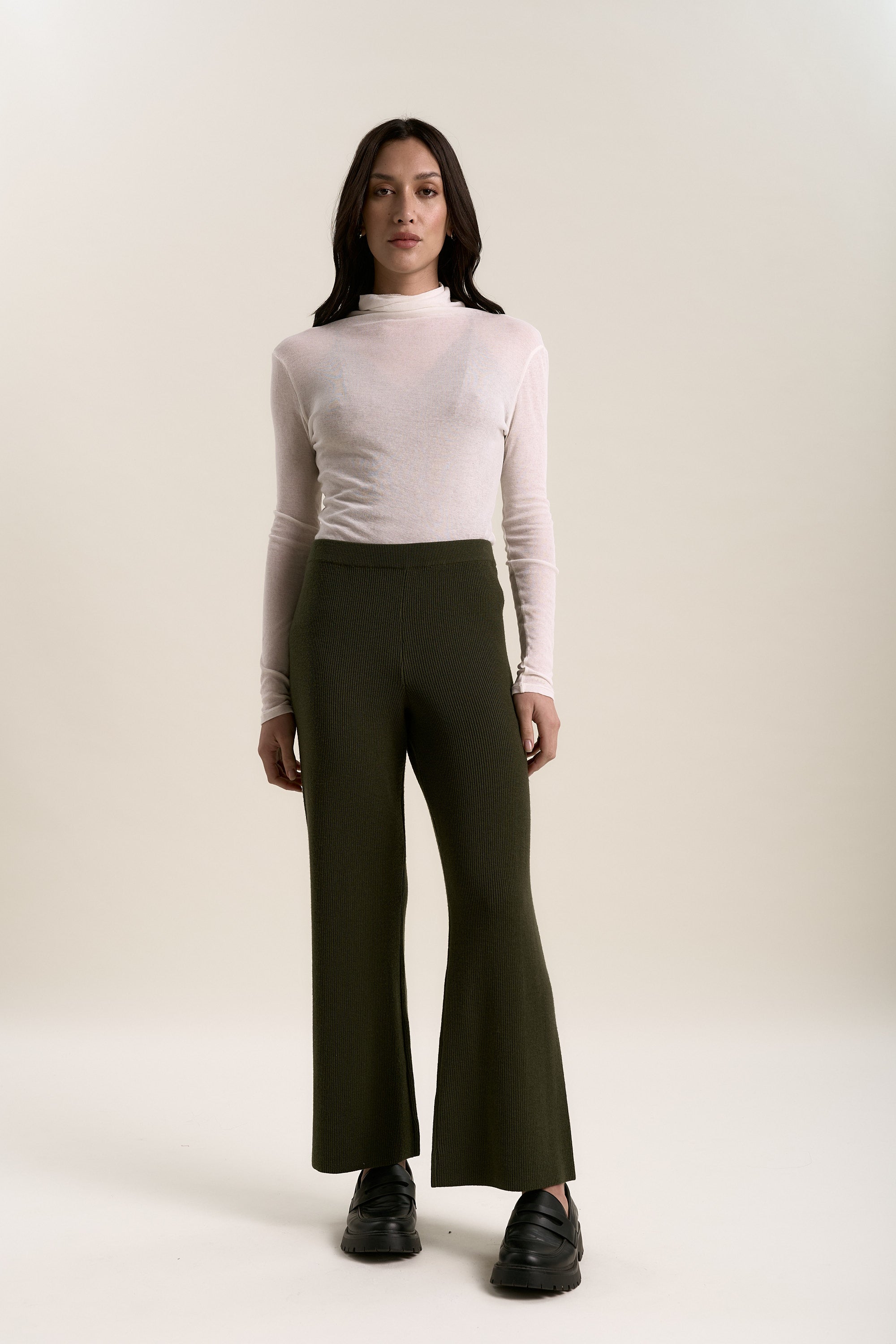 A person stands facing forward against a plain light background, wearing a sheer white turtleneck, black platform shoes, and Elementary Merino Blend Wide Leg Jogger pants by Elementary in olive green.