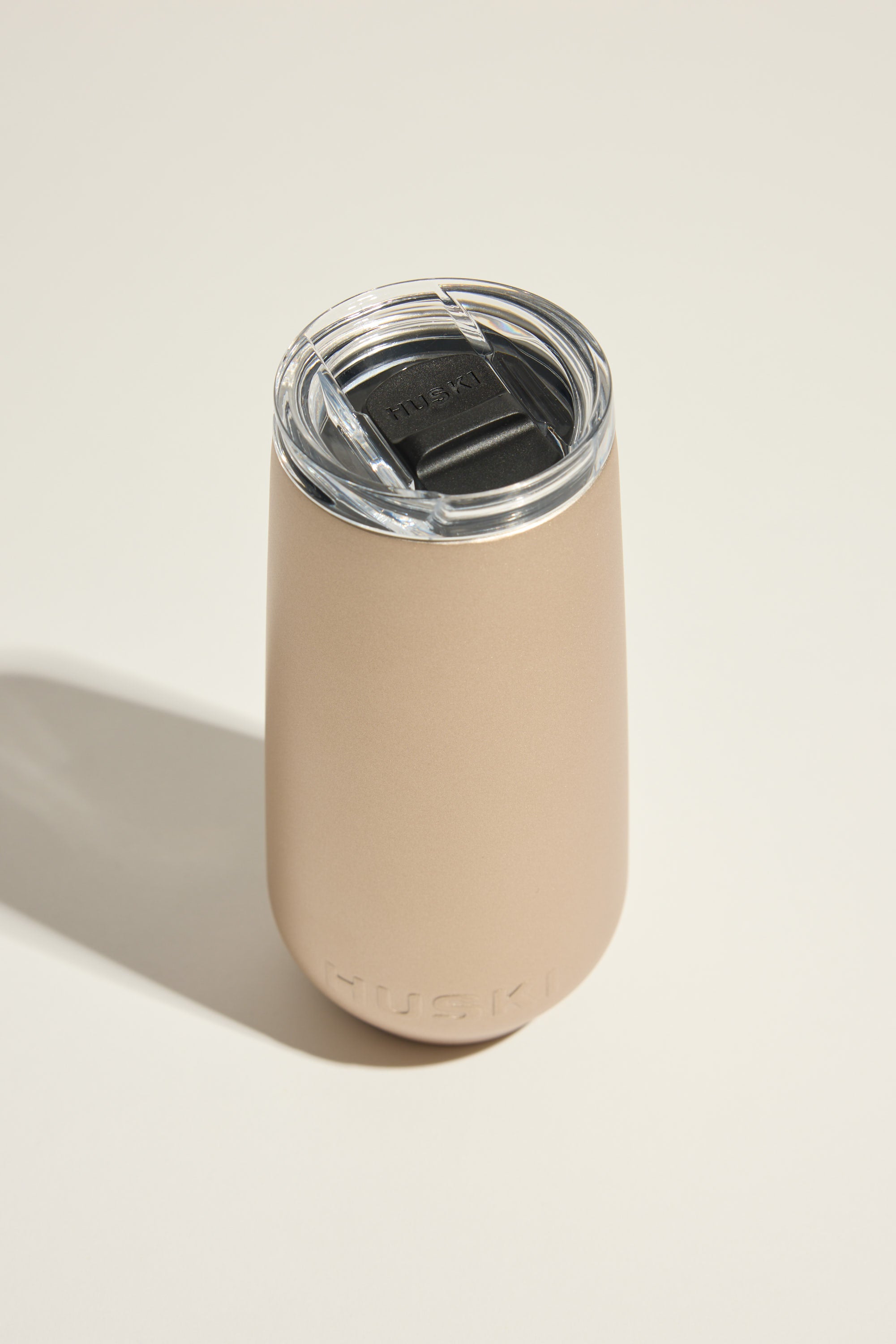 The Huski Champagne Flute by Huski is an insulated, beige travel tumbler with a clear plastic lid and black sliding closure. The word "Huski" is embossed near the base. Photographed on a light surface with a soft left shadow.
