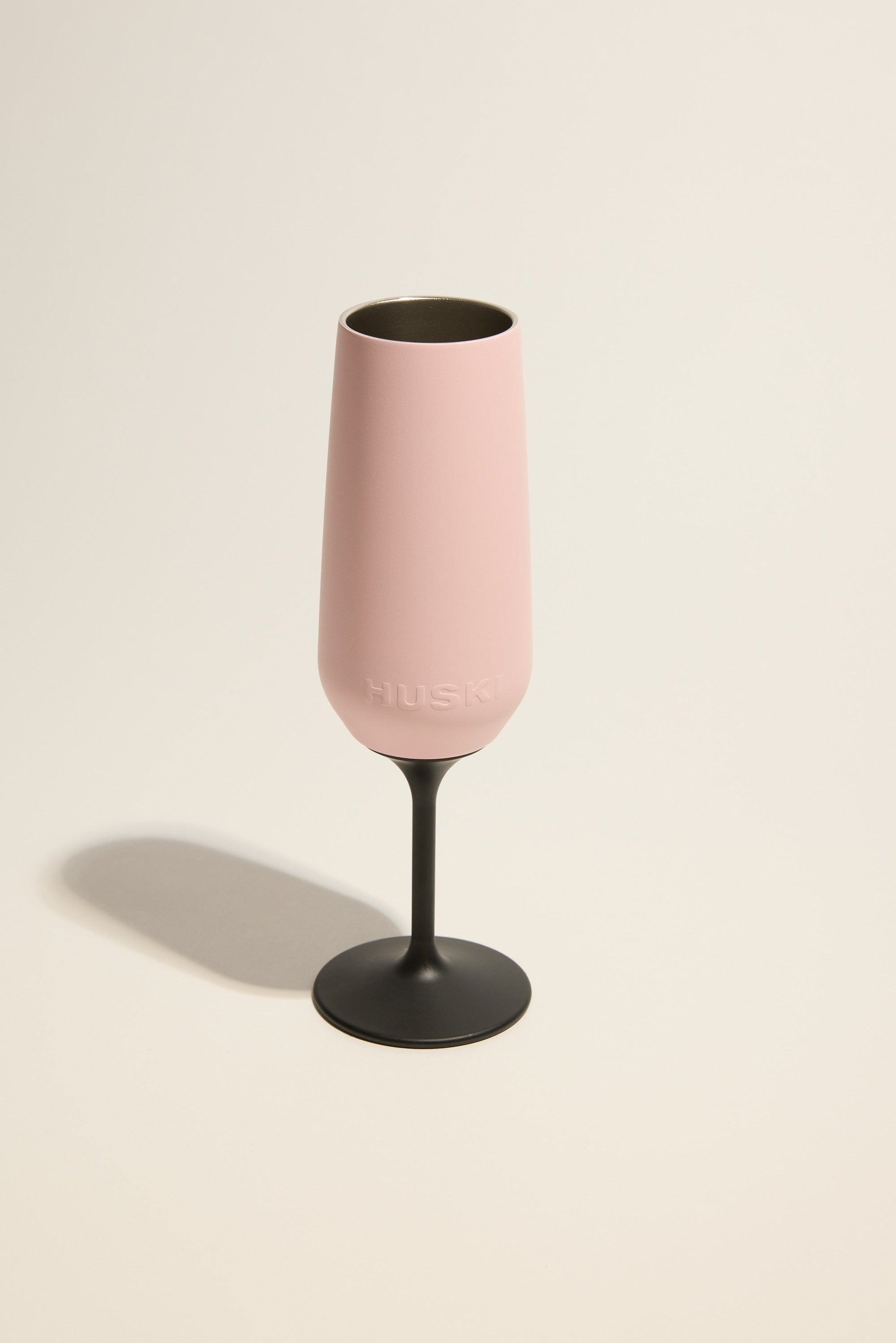 A Huski Champagne Flute with a pink cup and black stem stands on a light beige surface, casting a soft shadow. “HUSKI” is embossed on this insulated champagne flute from Huski.