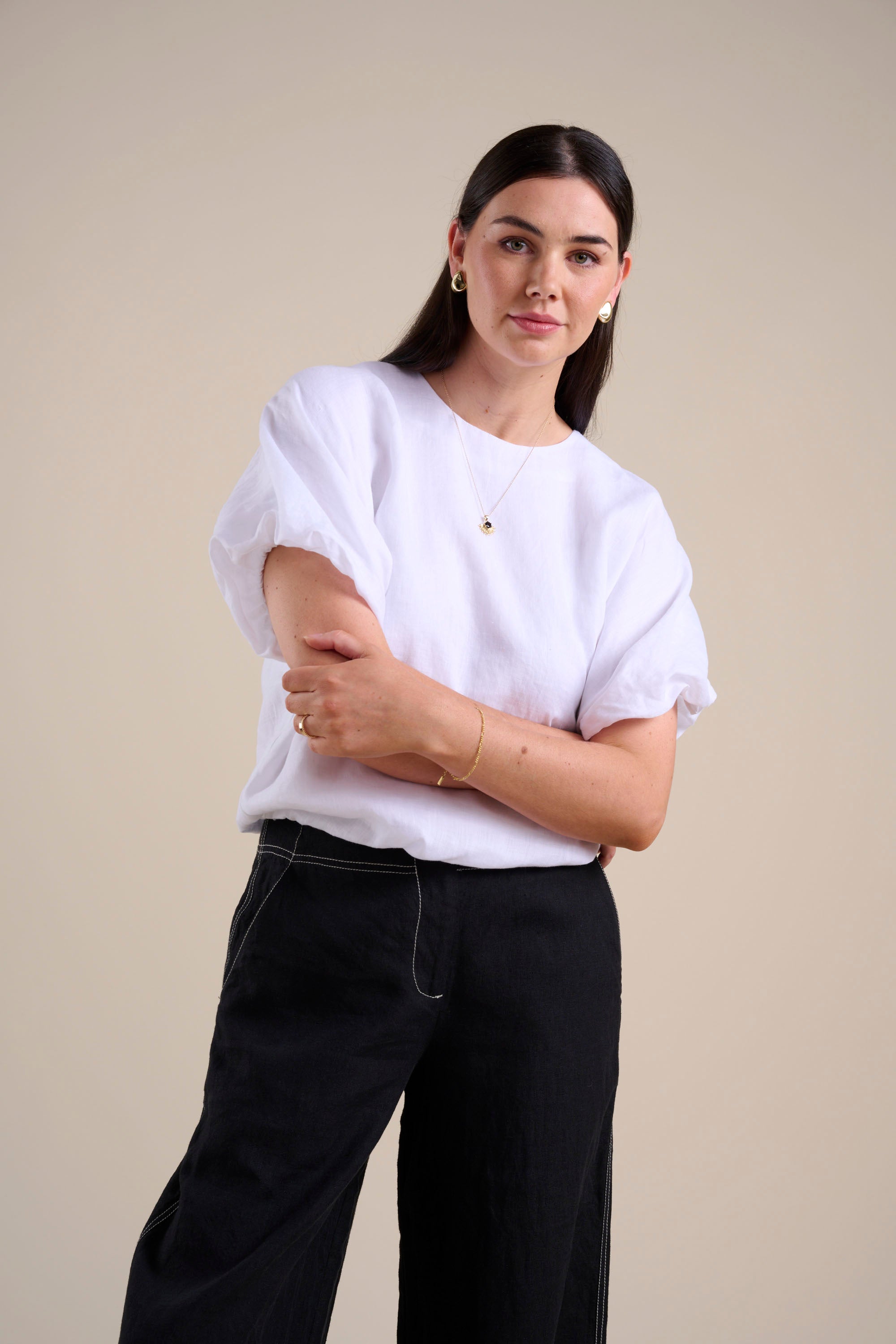 A woman with long dark hair stands against a plain background, wearing Max's The Bubble Hem Top with black pants and gold jewelry. She crosses her arms and gazes confidently at the camera.