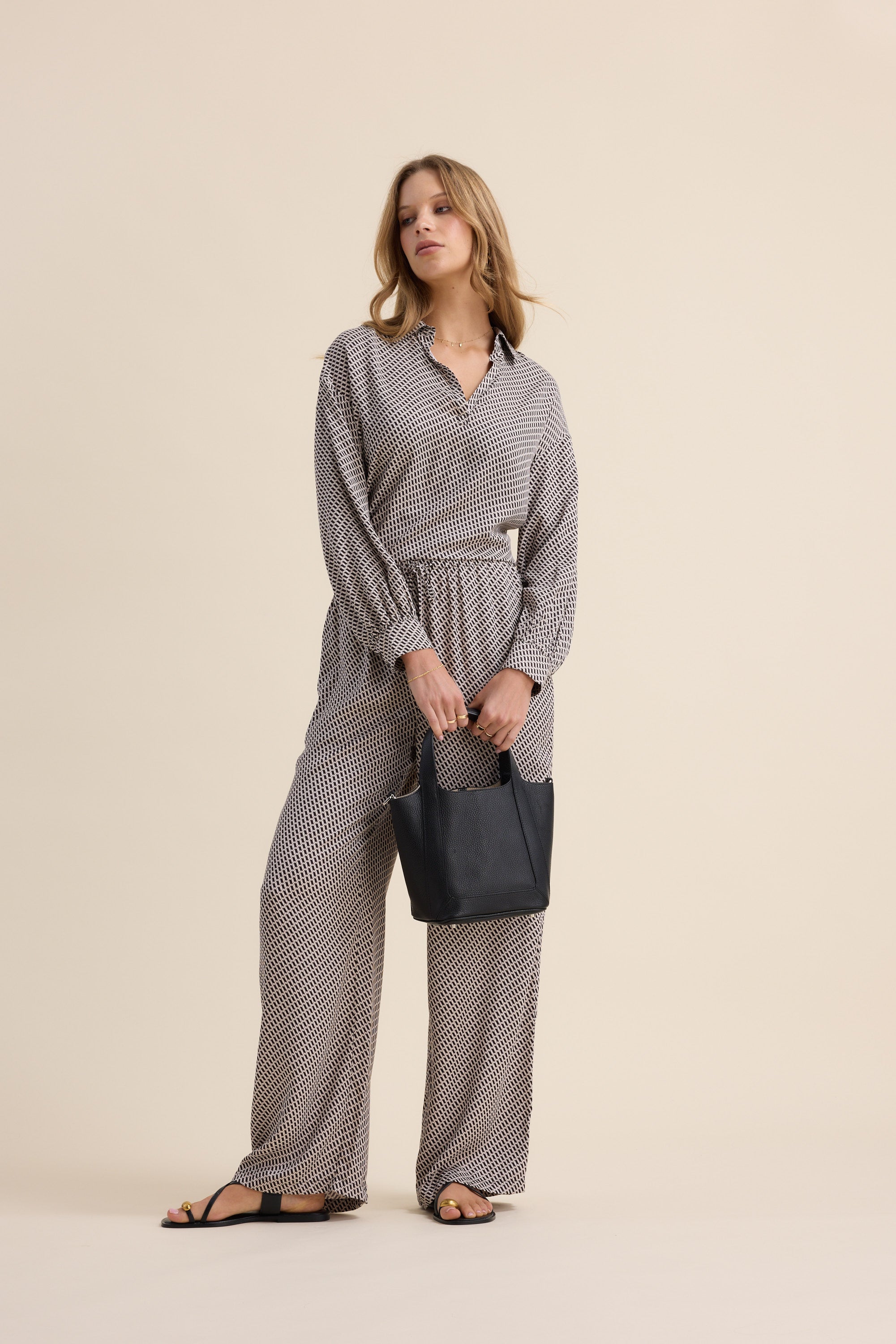 Woman in Max's The Luxe Pants holds a black handbag against a beige background.