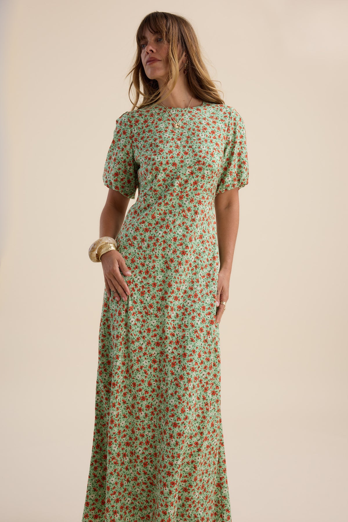 Woman wears Max’s Poppy Short Sleeved Dress, green floral, against a plain background.