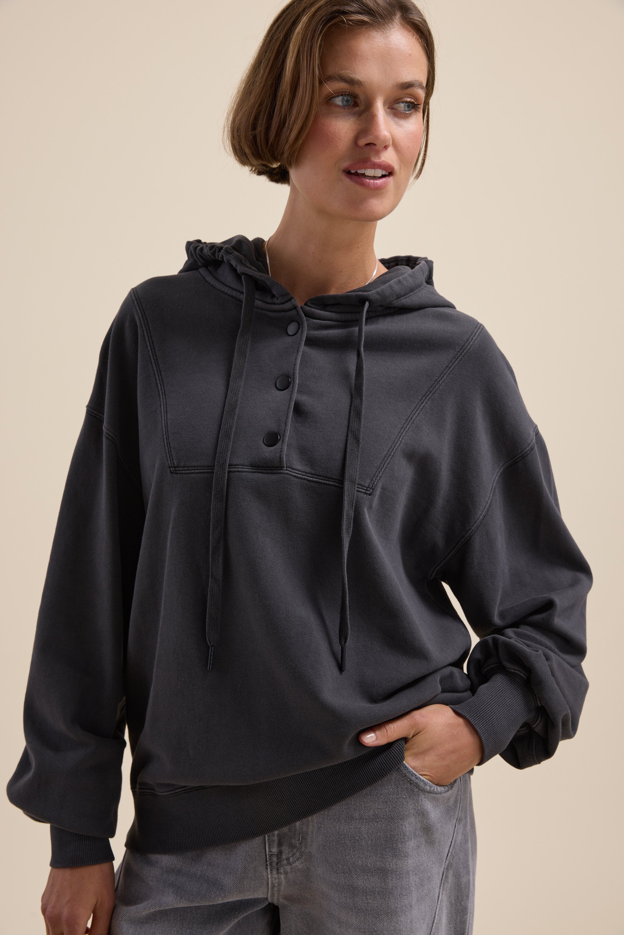 Woman in an Elementary Henley Hoodie and light gray pants, hand in pocket.