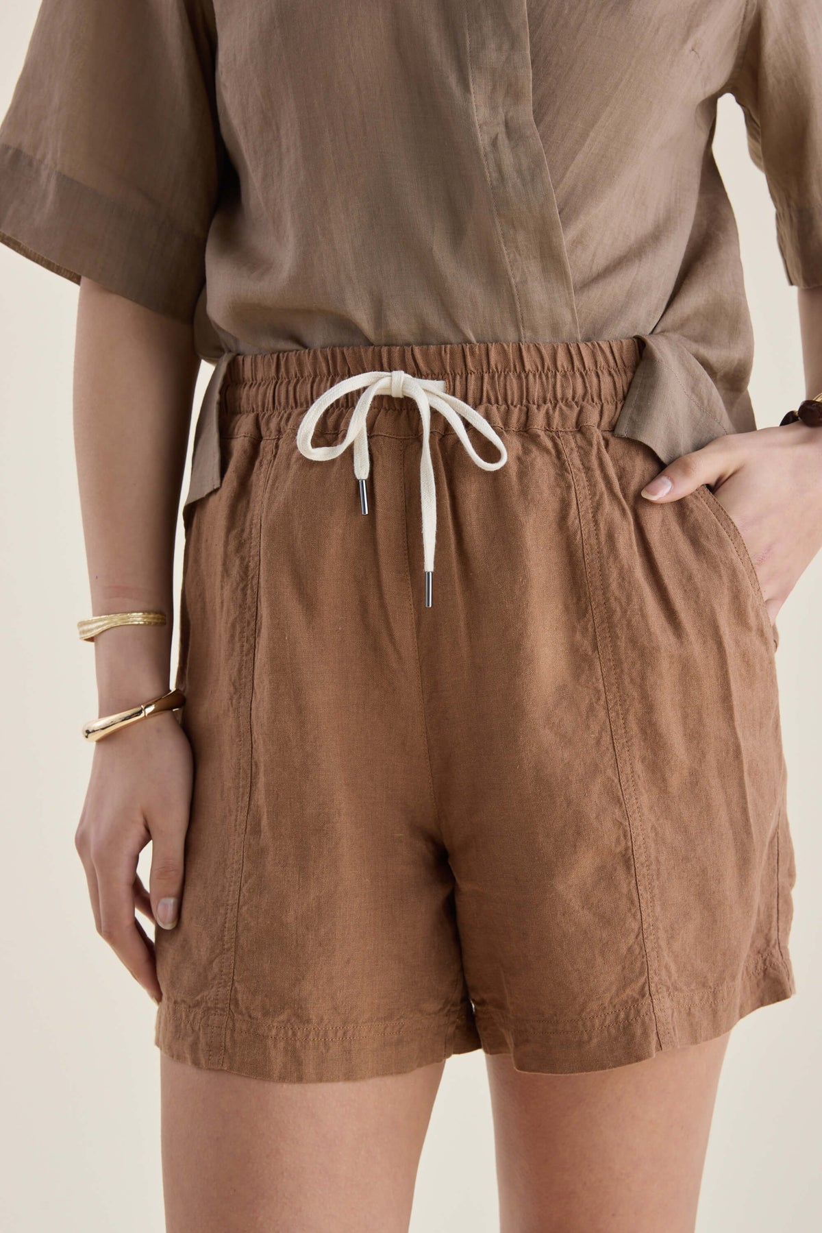 Model in Elementary’s high-rise linen shorts with drawstring, brown shirt, and gold bangles.
