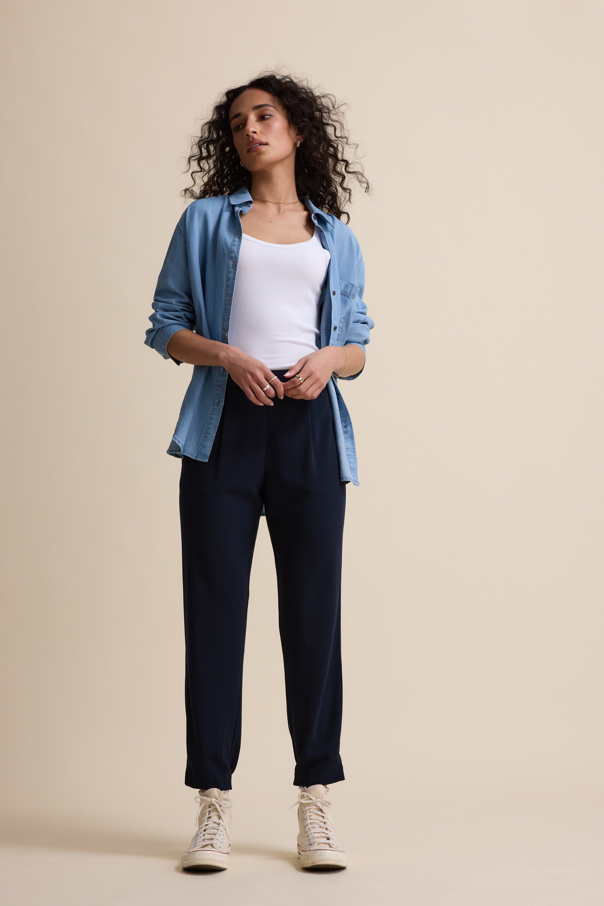 A woman with curly hair stands against a beige background, wearing Max’s The Easy Care Ankle Grazer, a light blue button-up over a white tank, and light high-top sneakers. One hand in her pocket, she looks confidently ahead.