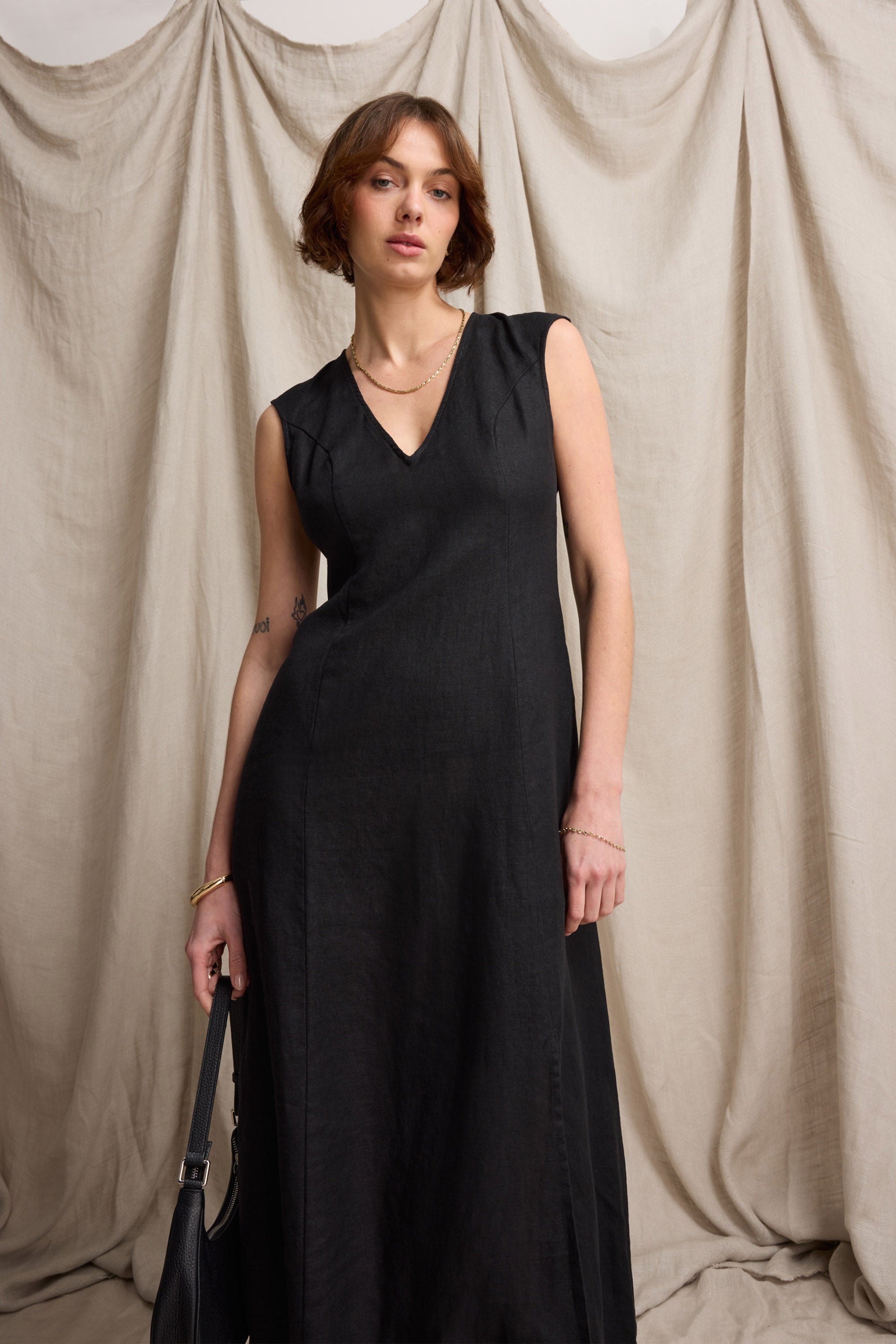 Woman wears Max's Alice V Neck Linen Dress, standing before beige draped fabric, holding a bag.