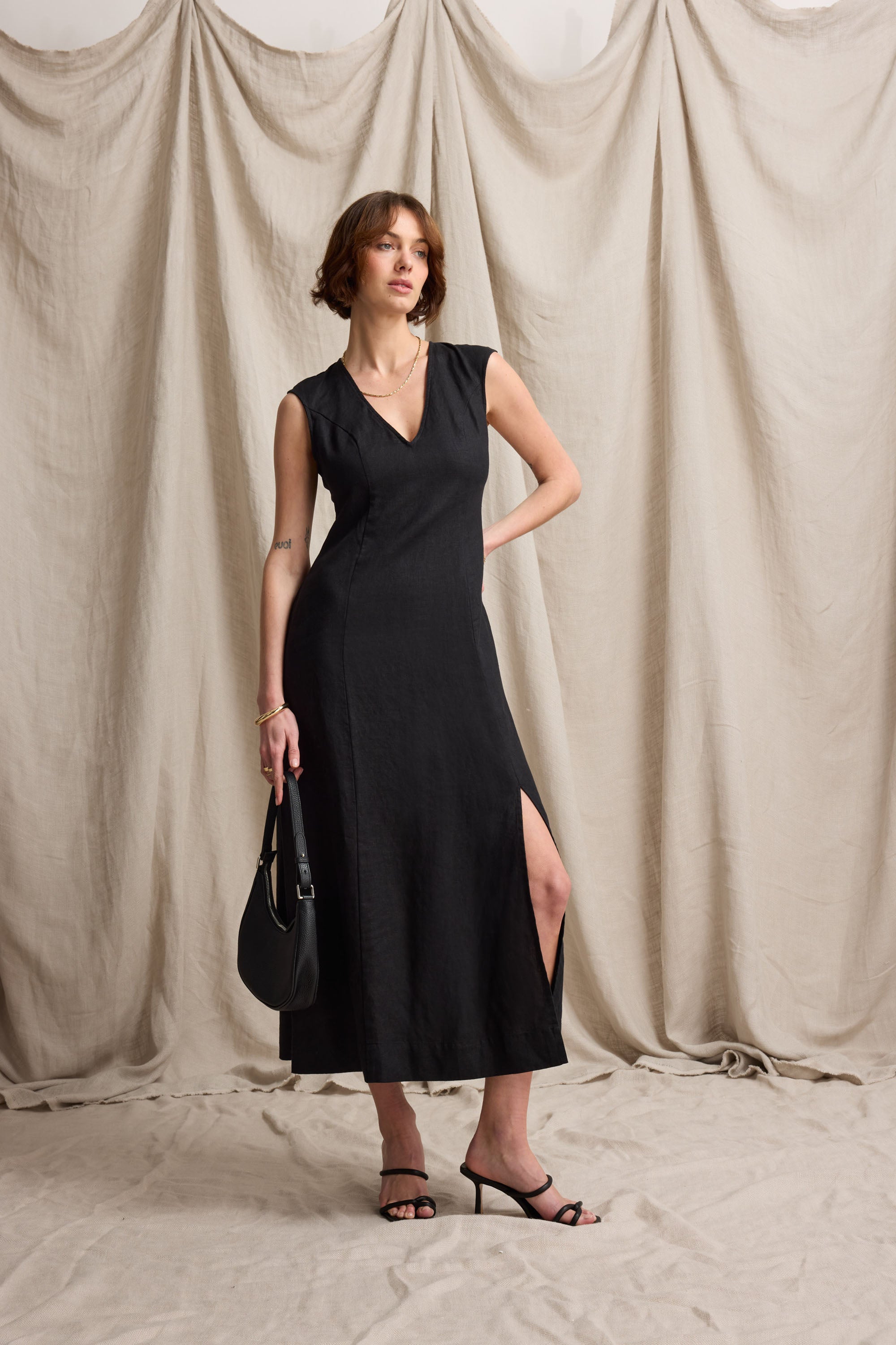 Woman in the Max Alice V Neck Linen Dress and heels poses with a bag before beige draped fabric.