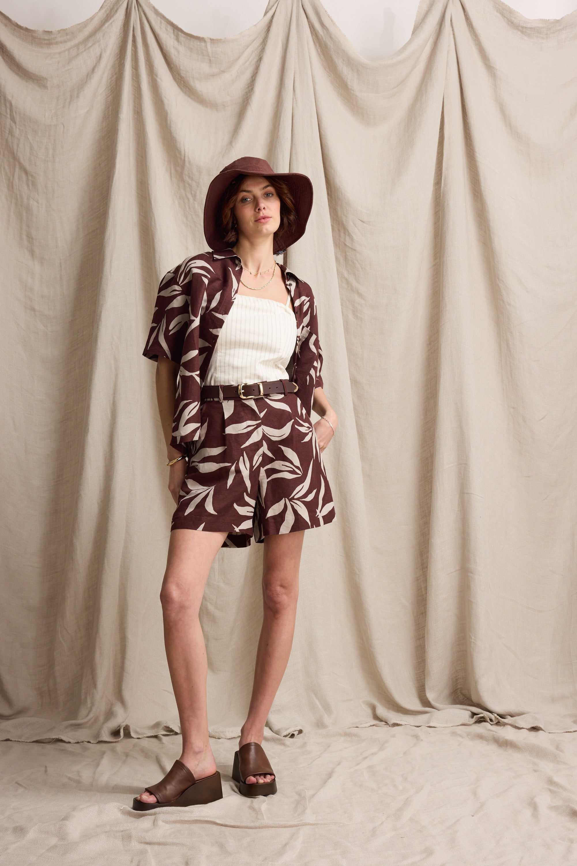 Woman poses in Max Claire Linen Blend Pleated Short, hat, and sandals against a beige backdrop.