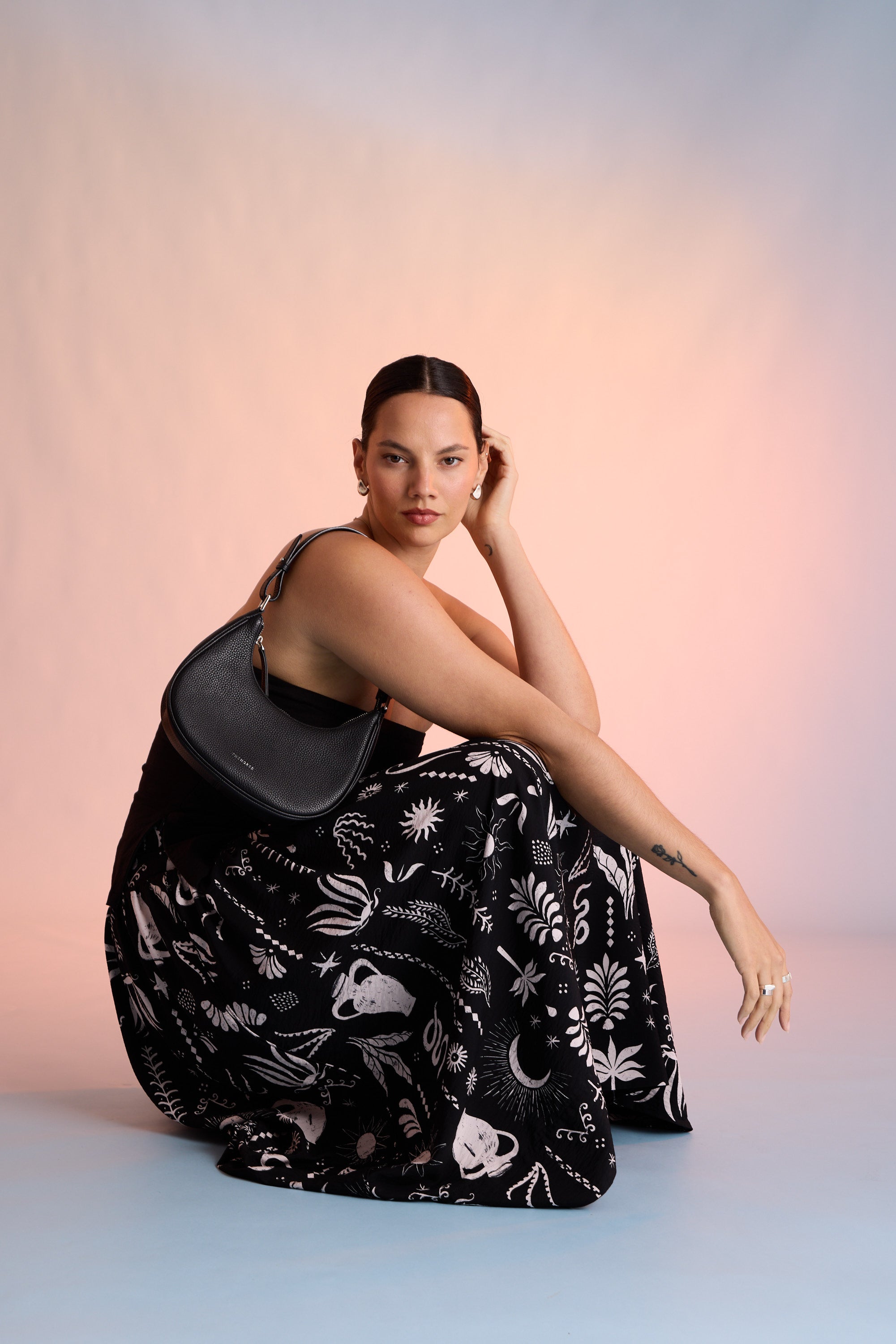 A woman with dark hair poses sideways, wearing the Max Helena Drop Waist Skirt with exclusive celestial and floral prints, paired with a black strapless top and holding a black shoulder bag against a pastel background.