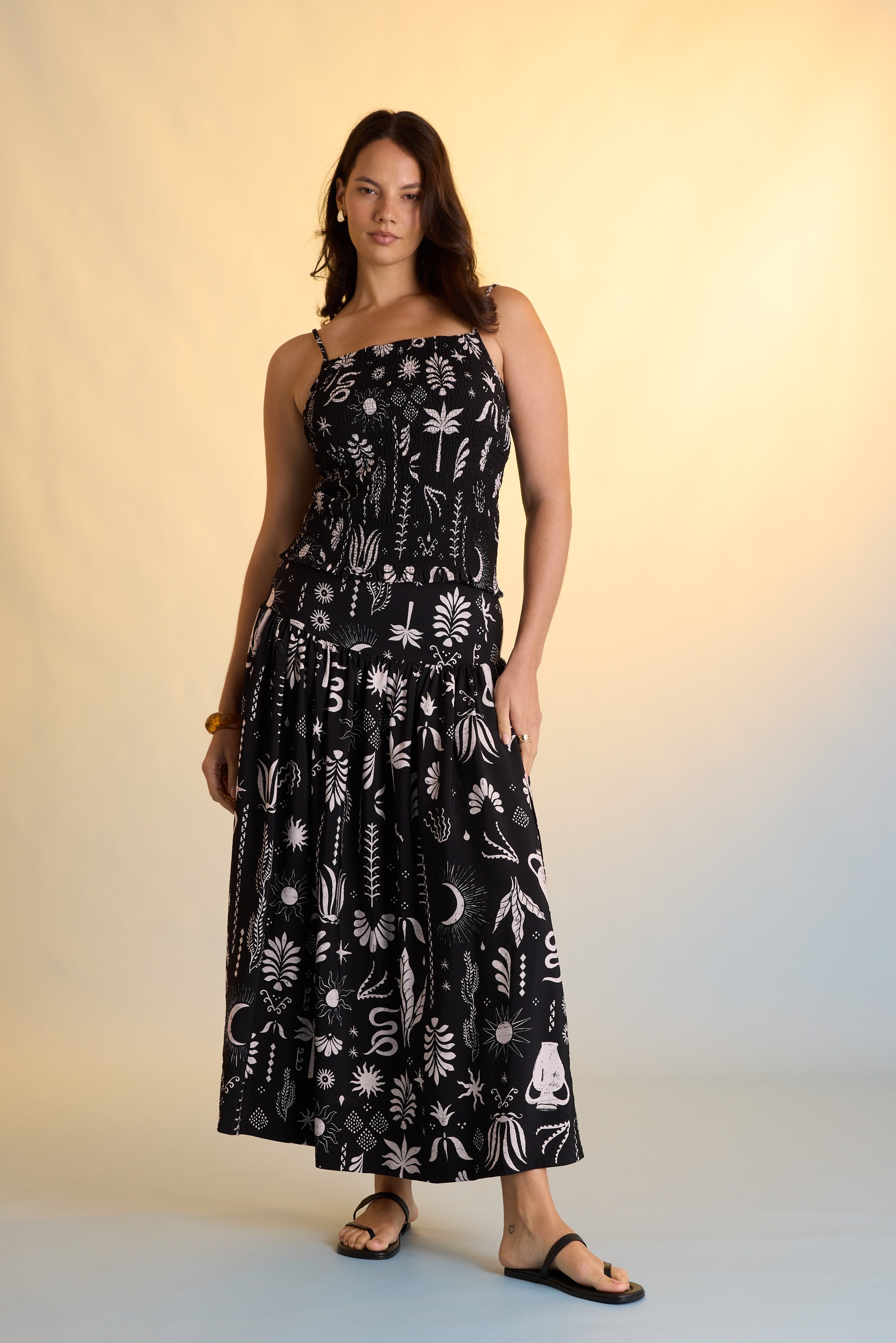A woman stands against a warm gradient background, wearing the Max Helena Drop Waist Skirt with exclusive black fabric featuring white botanical and celestial prints, paired with black sandals and minimal accessories.