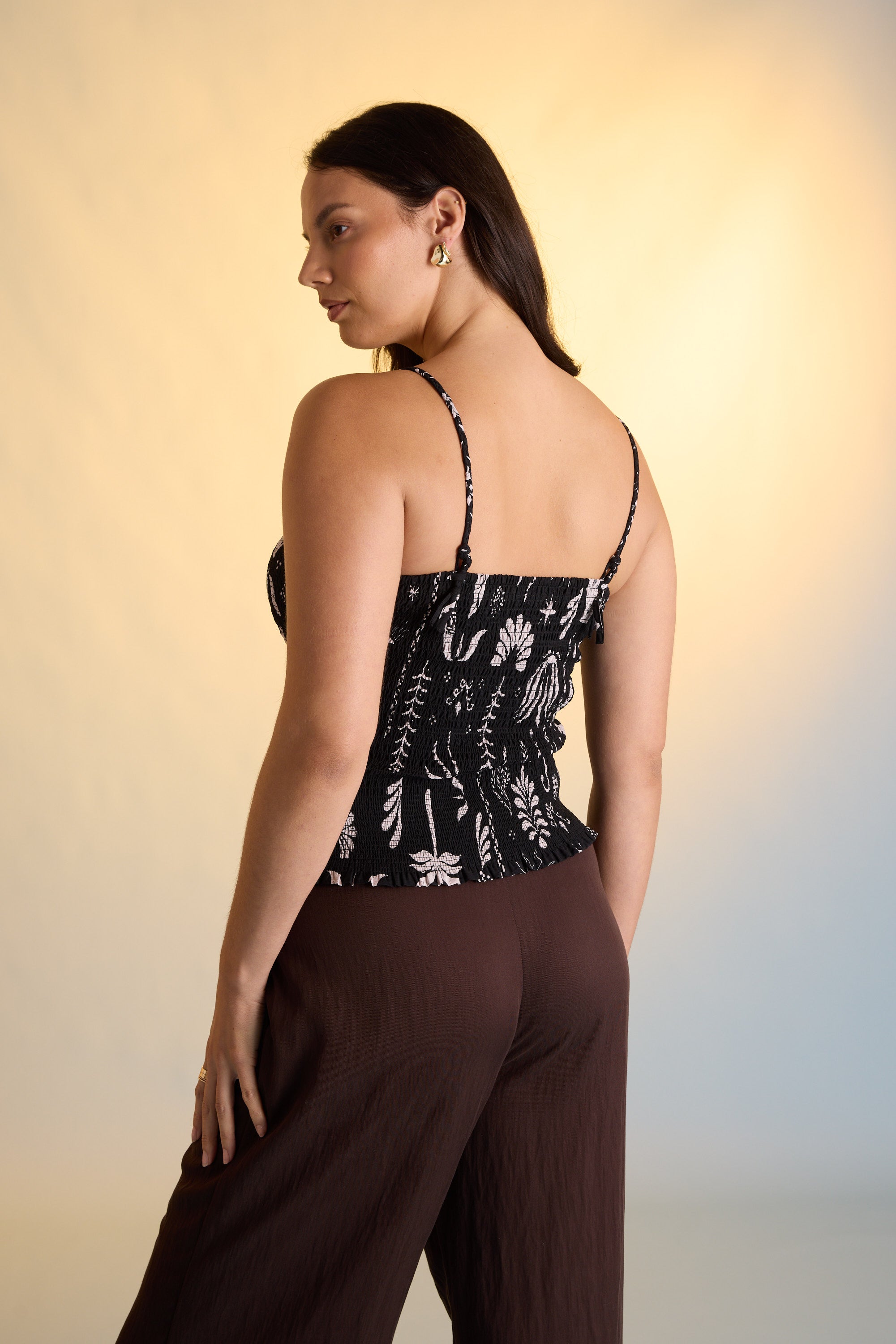 A woman with long dark hair wears the Max Emma Shirred Cami in black with a floral pattern and brown wide-leg pants, standing turned away and looking over her shoulder against a softly lit gradient background.