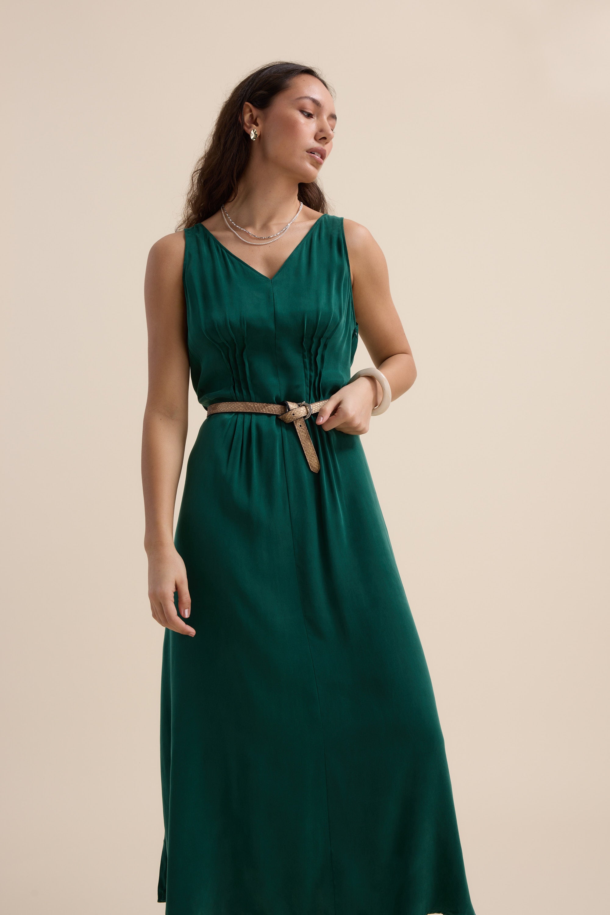 A woman models the Max Cara Pleat Detail Dress, a sleeveless emerald green V-neck with a tan belt, cinched waist, and pleats, styled with earrings, necklace, and bracelet against a plain beige background.