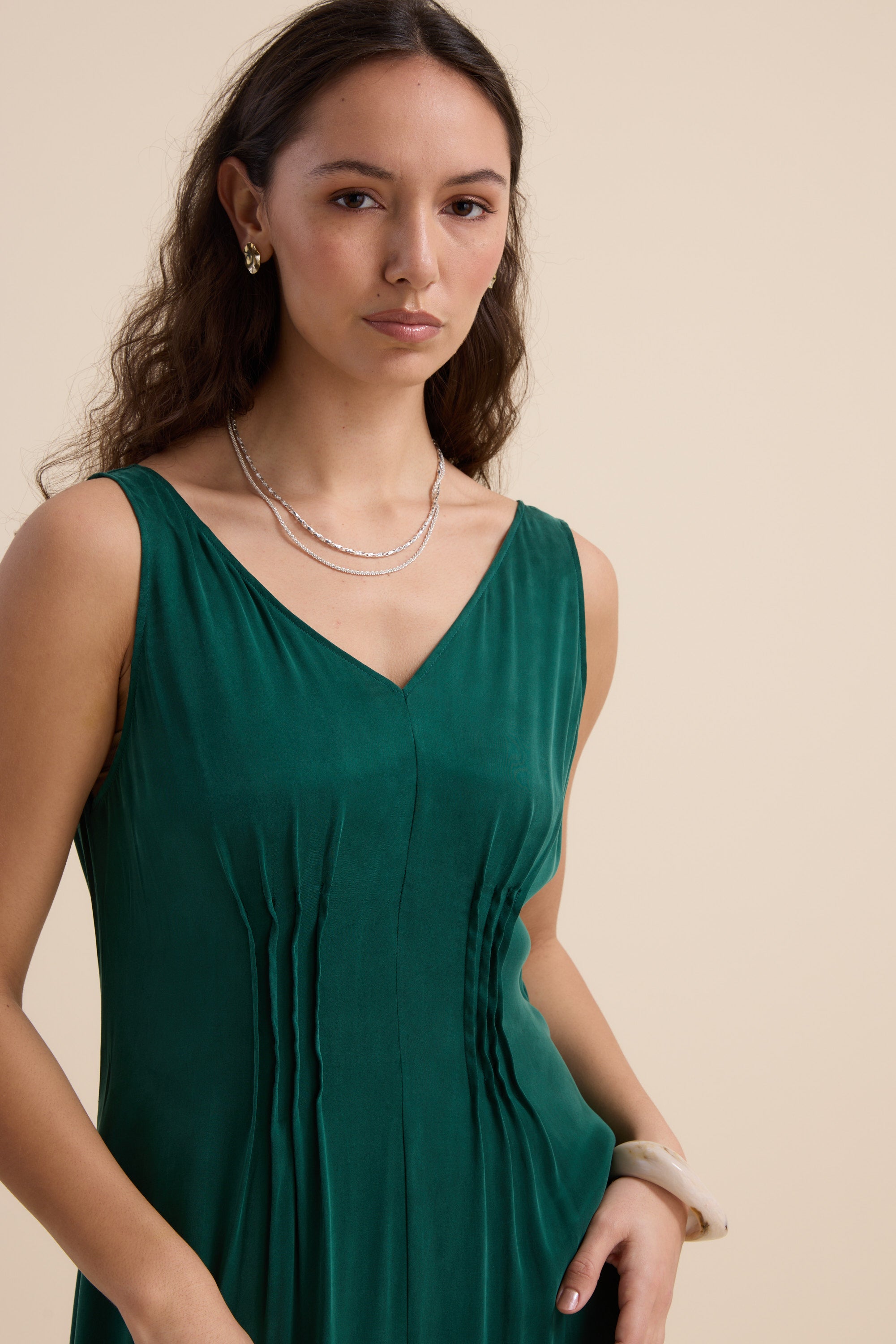 A woman with long brown hair wears the Max Cara Pleat Detail Dress, an emerald green sleeveless dress with a cinched waist, paired with a double-strand necklace, earrings, and bracelet, as she poses confidently against a beige background.
