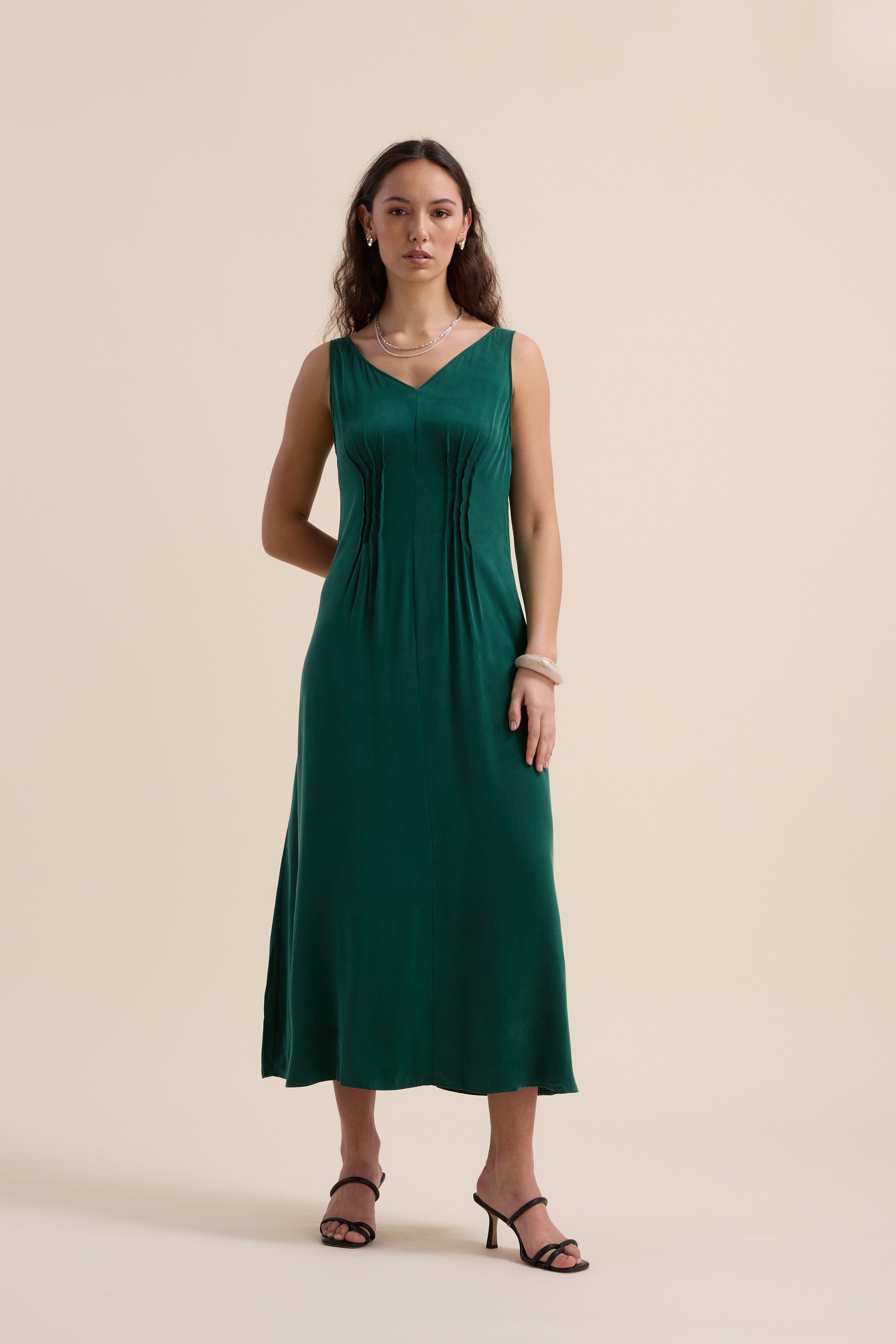 A woman wears the Max Cara Pleat Detail Dress, a sleeveless emerald green maxi with pleats, cinched waist, and V-neckline. She pairs it with bracelets, a necklace, and black heeled sandals against a beige background.